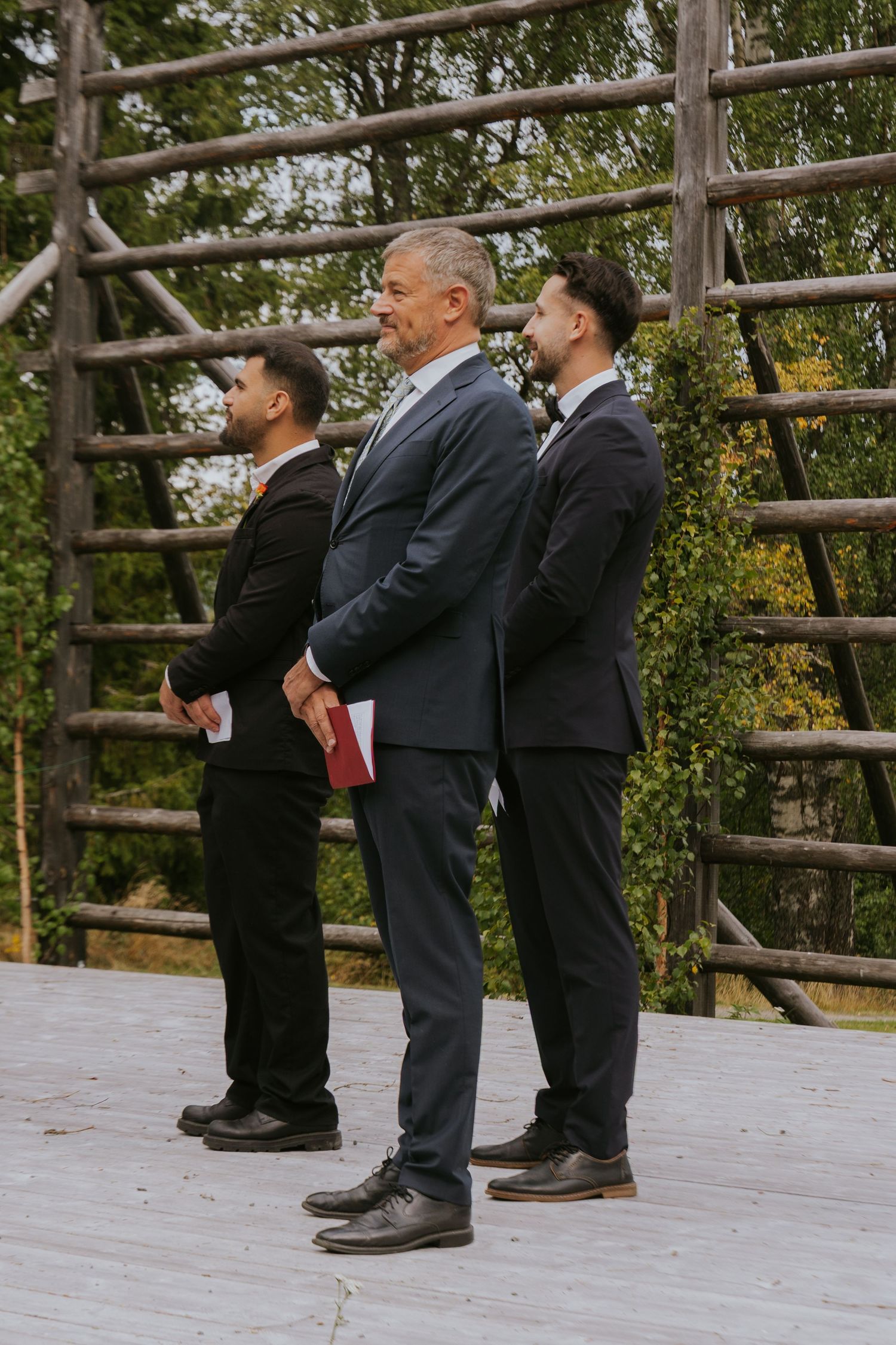 Groomsmen in formal attire stand in line on a wooden deck during an outdoor wedding ceremony.