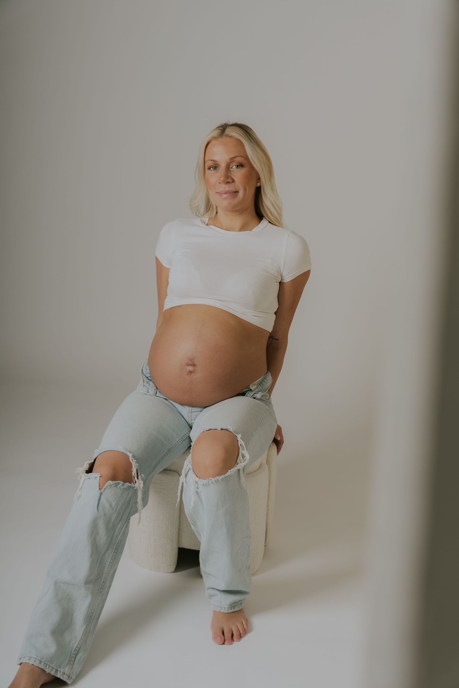 Maternity photo of an expectant mother in distressed jeans and white crop top against neutral background.