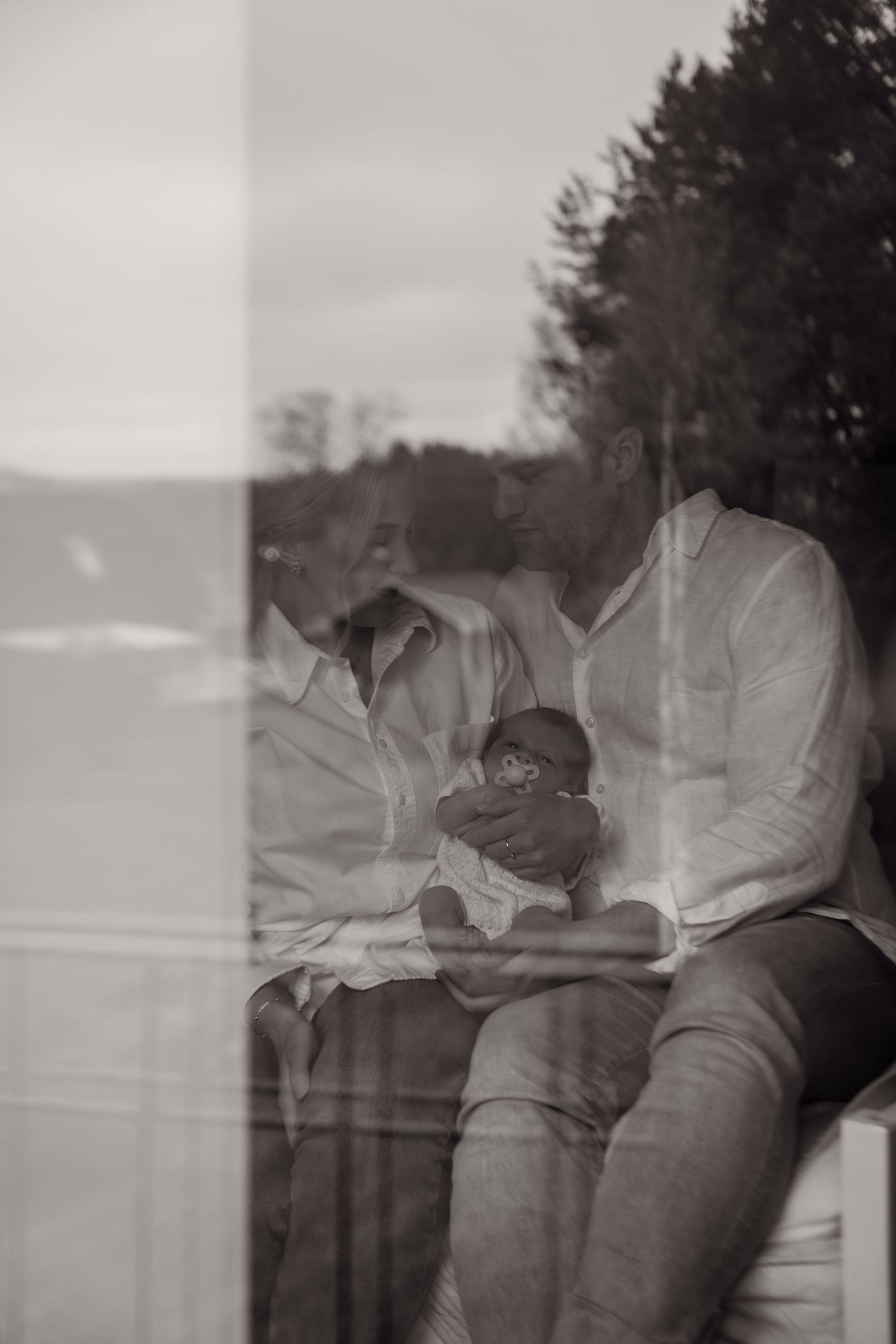 A black and white artistic photograph showing a person reading a book through a window reflection.