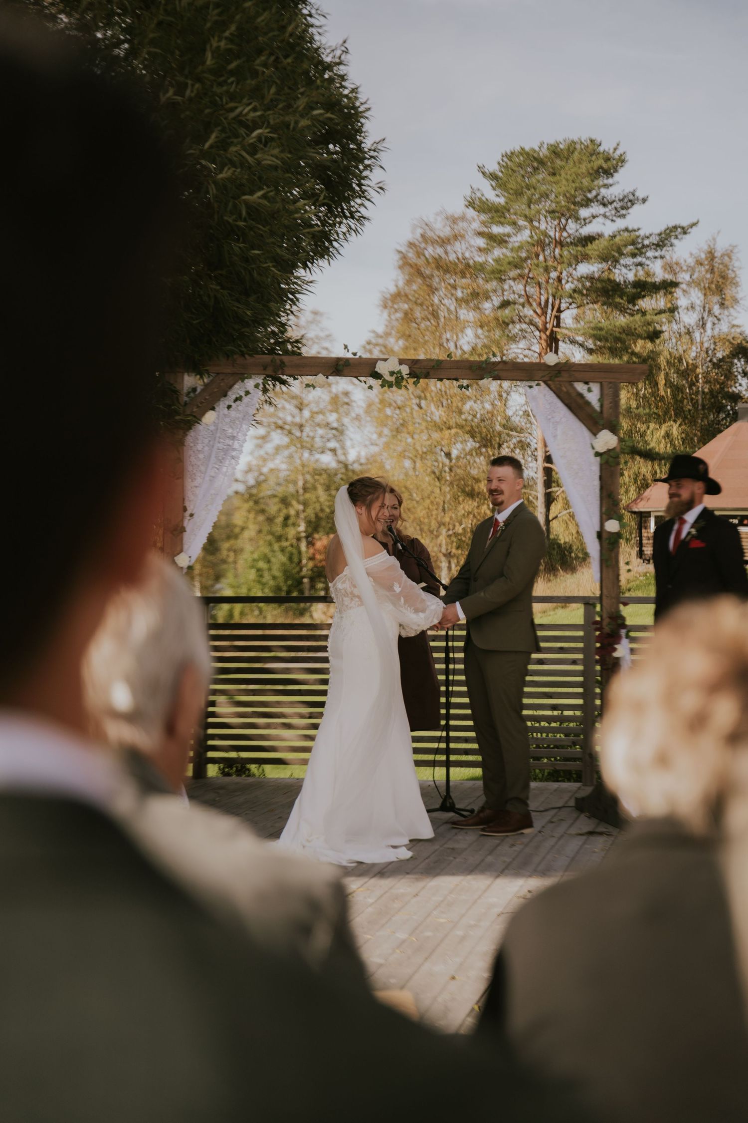Wedding ceremony under wooden pergola with guests watching from their seats.