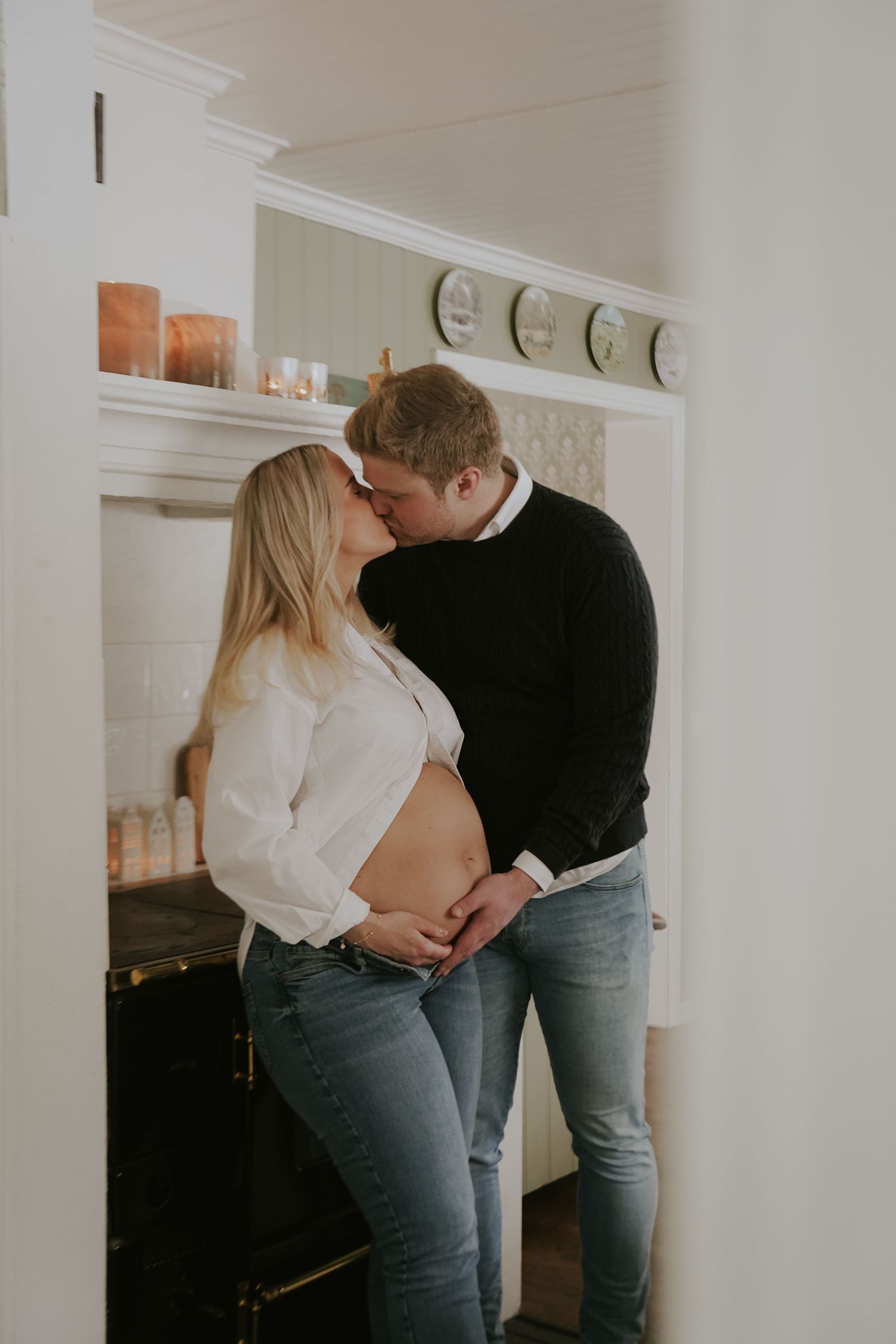 Romantic couple in casual attire embracing in a minimalist white room with soft lighting.