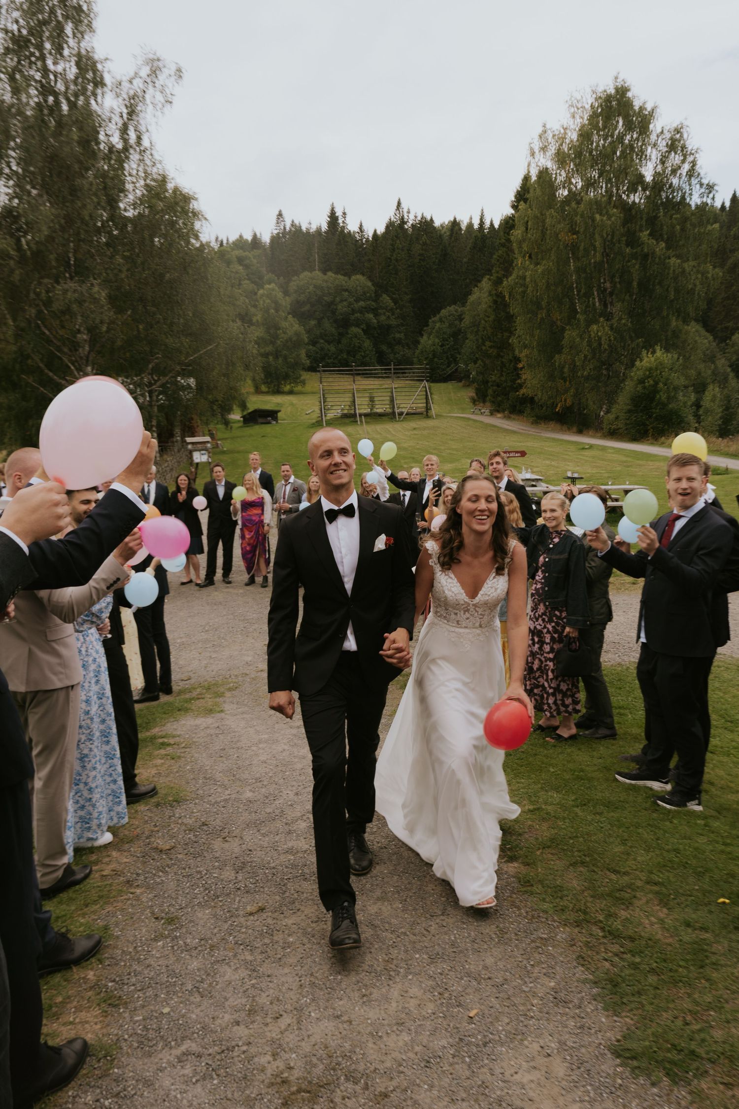A couple walks down a path lined with wedding guests holding pink and white balloons in a wooded outdoor setting.