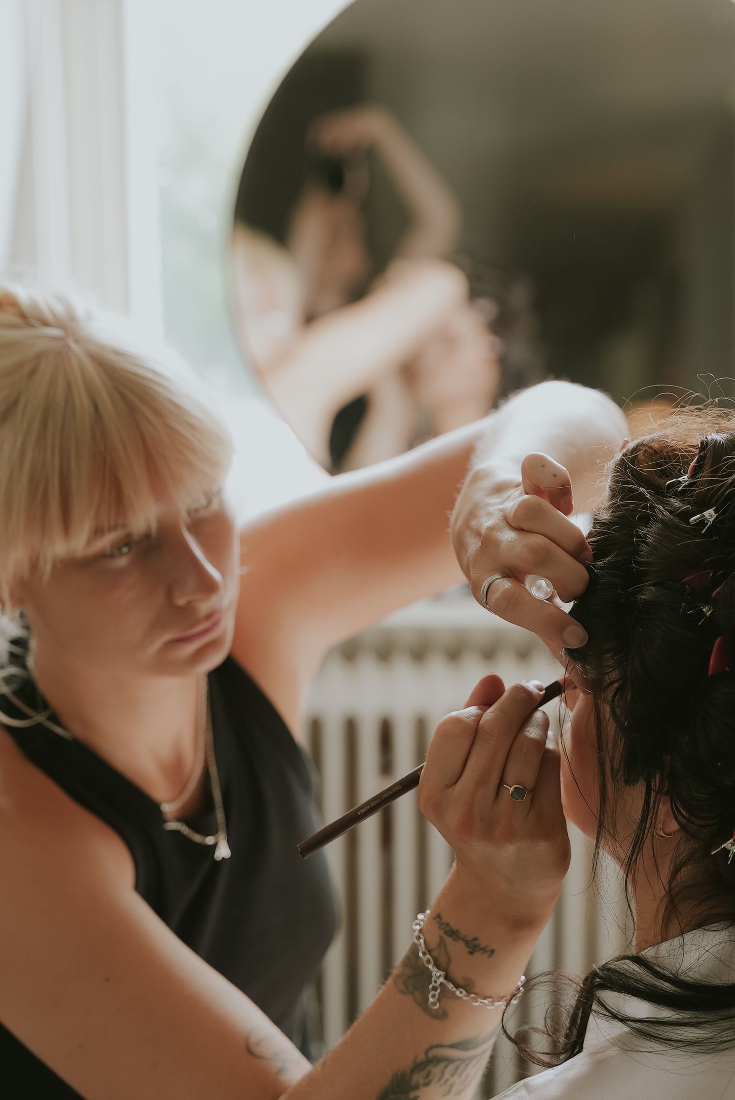 Hair stylist working on client's updo hairstyle in soft natural lighting during wedding preparation.
