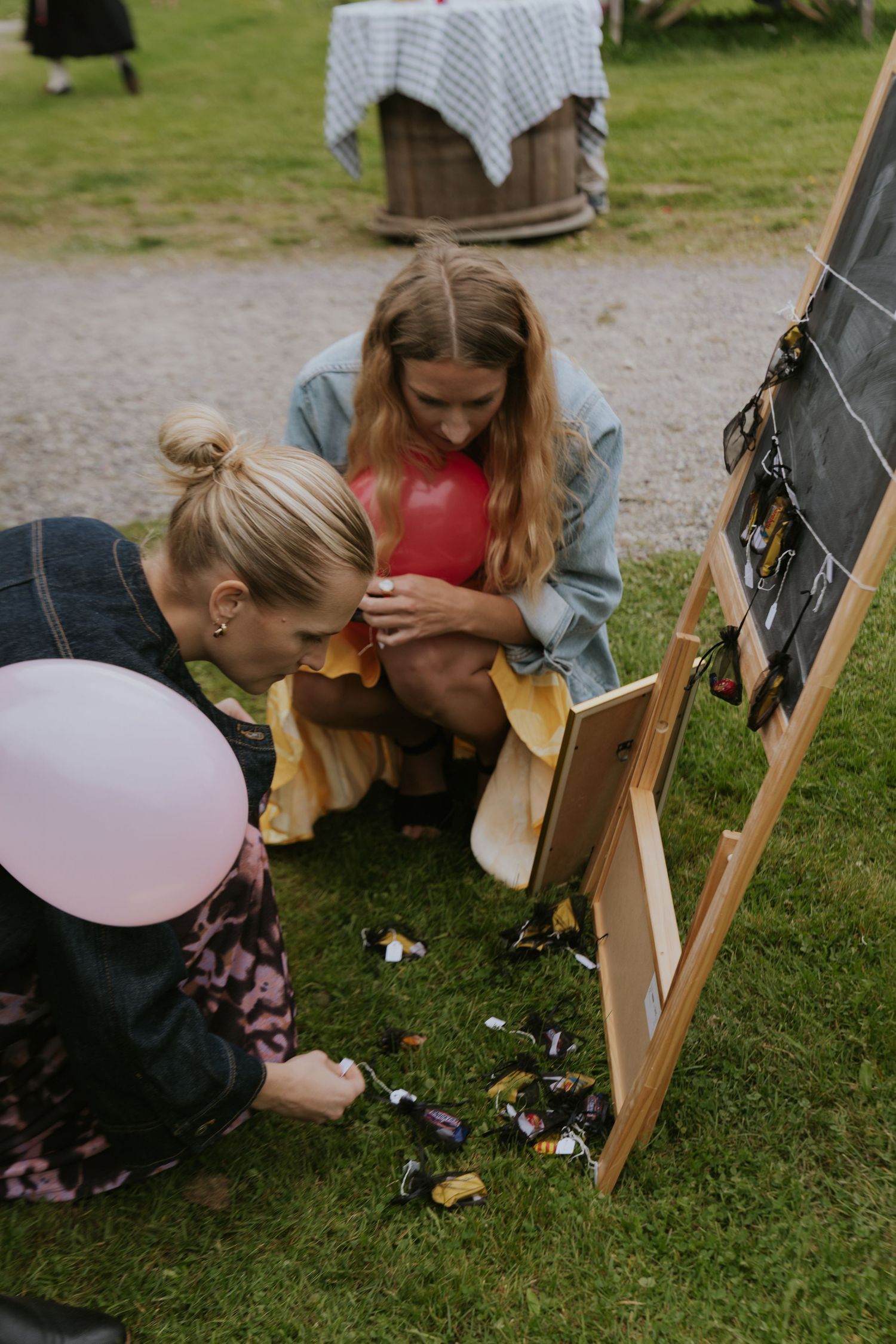 Two people sitting on grass with balloons and a chalkboard at an outdoor party setting.