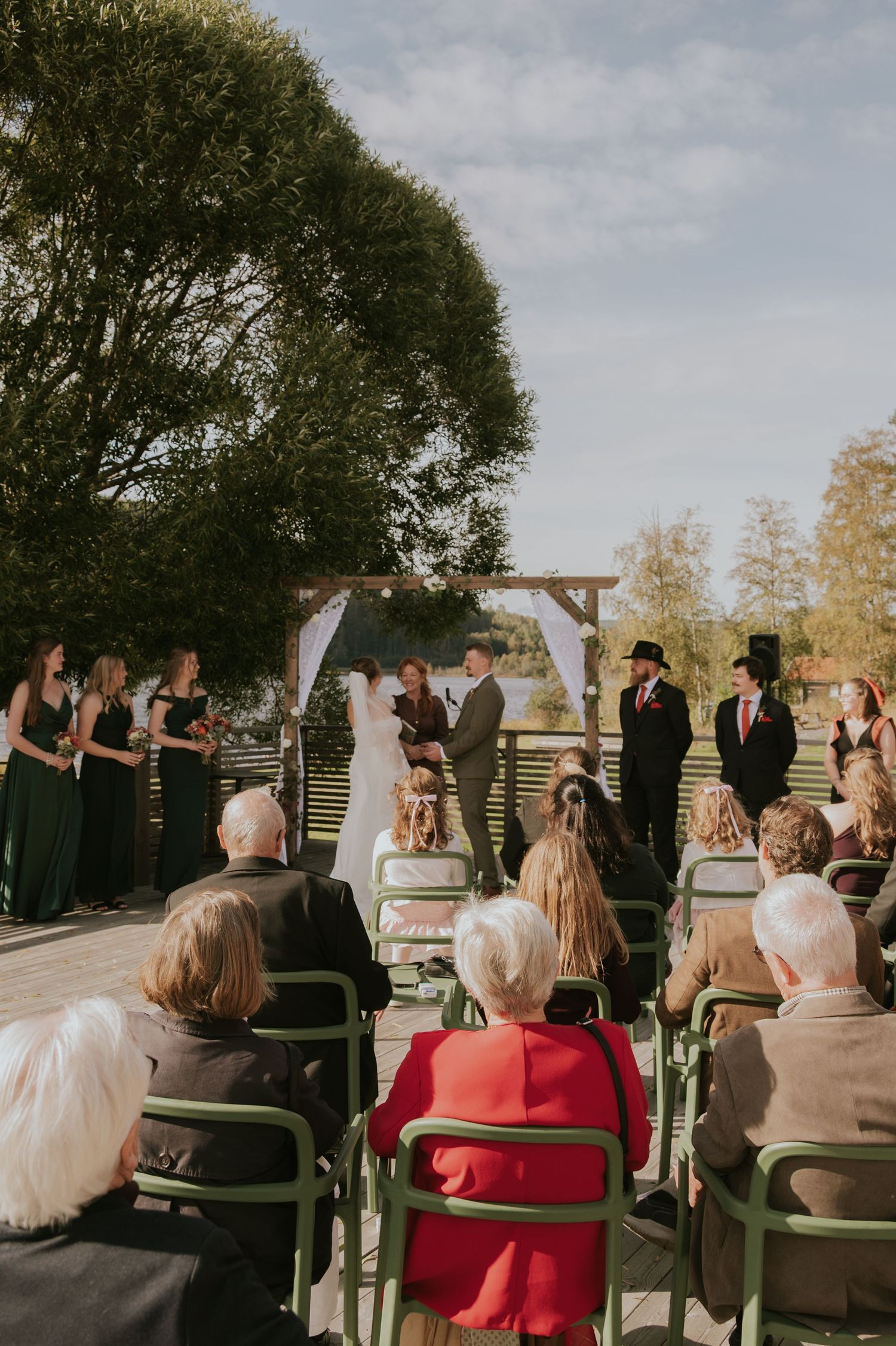 Wedding guests seated in green chairs watch an outdoor ceremony under trees and cloudy skies.