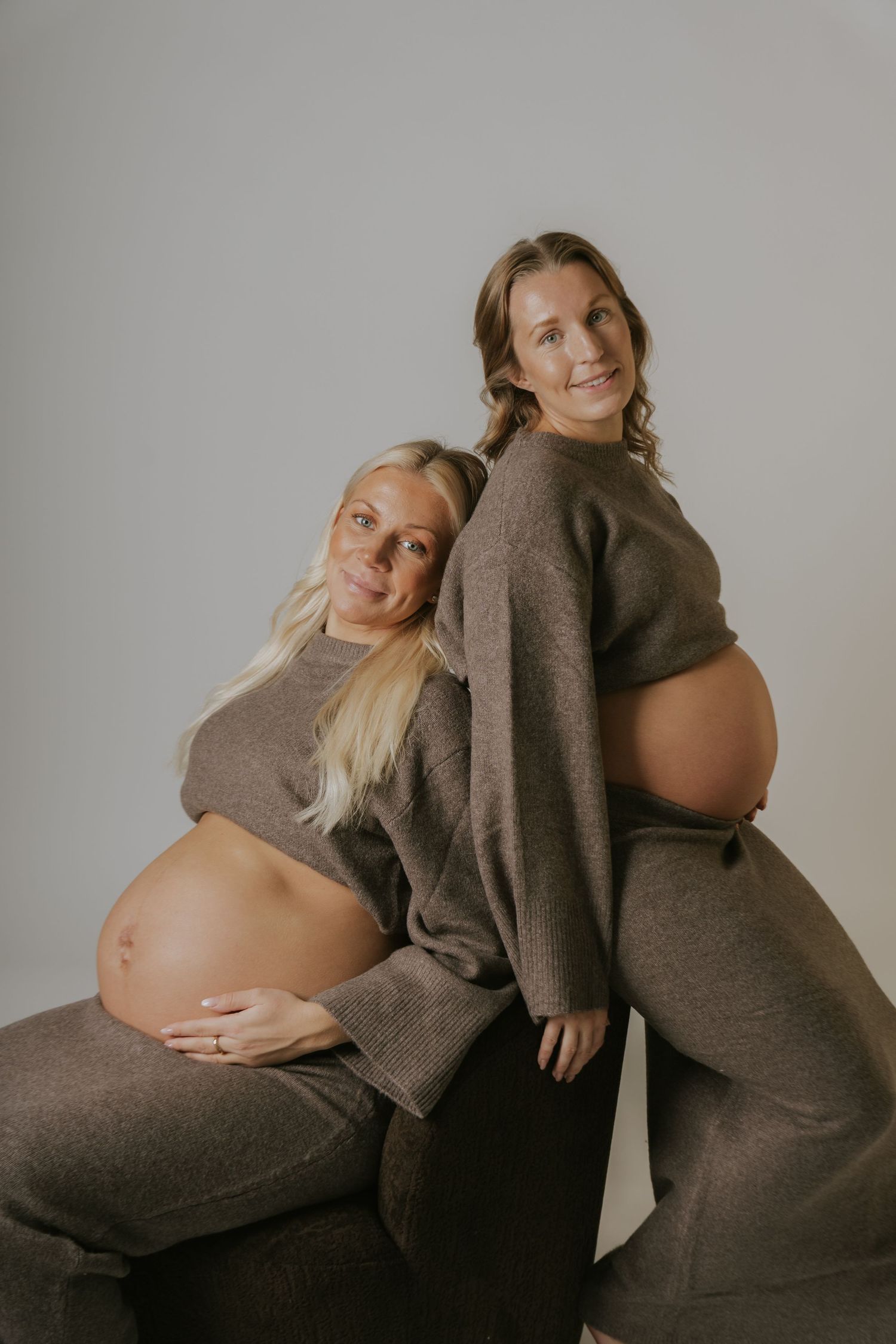 Two pregnant women in brown sweaters embracing and posing together.