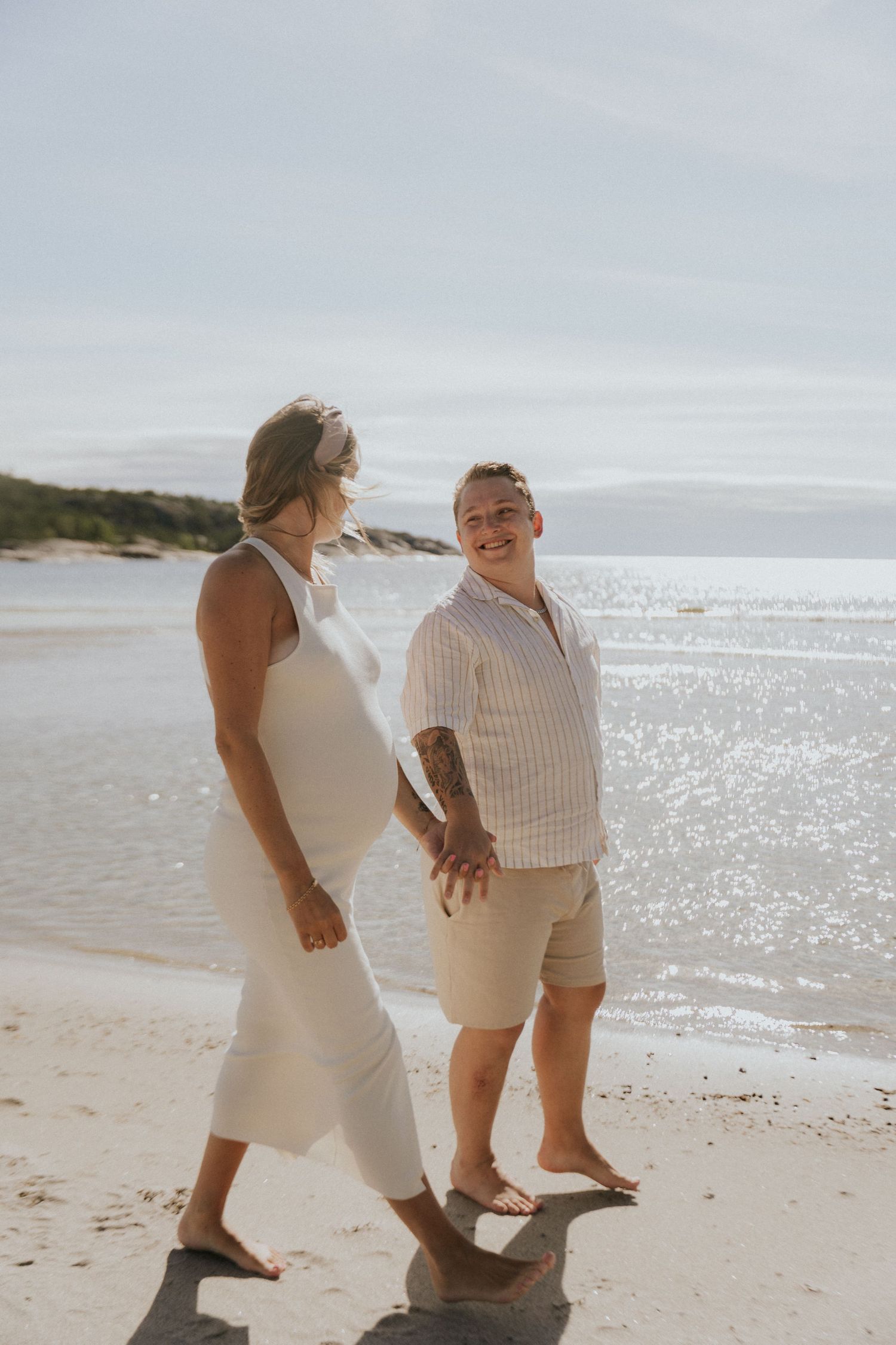 Couple sharing playful moment on sunny beach during maternity photoshoot.