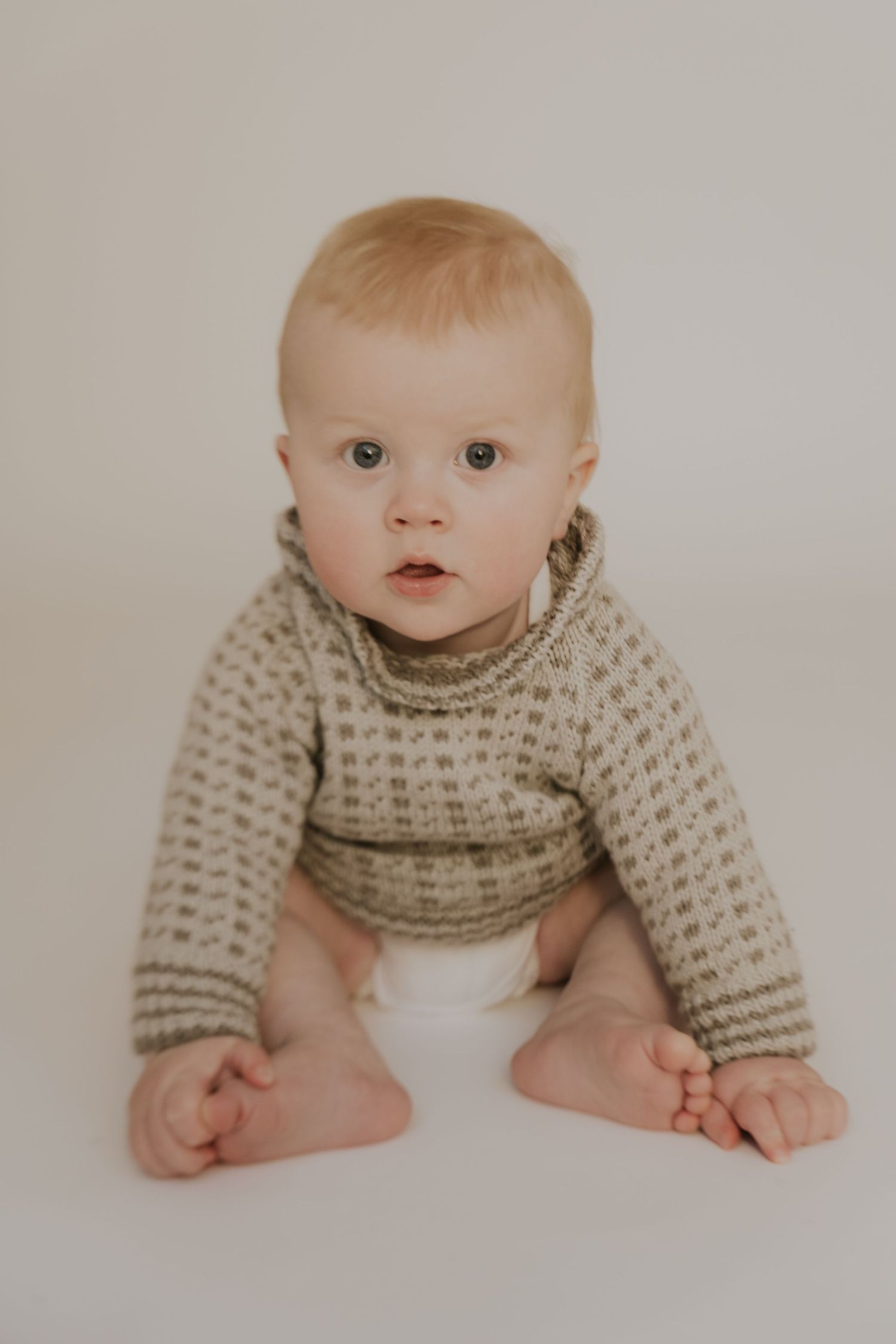 Baby wearing knit sweater sitting on white background looking at camera.