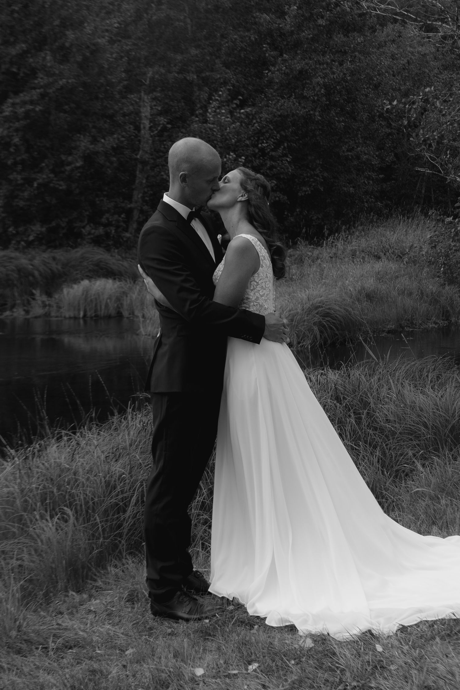 Black and white wedding portrait of couple kissing by waterside.