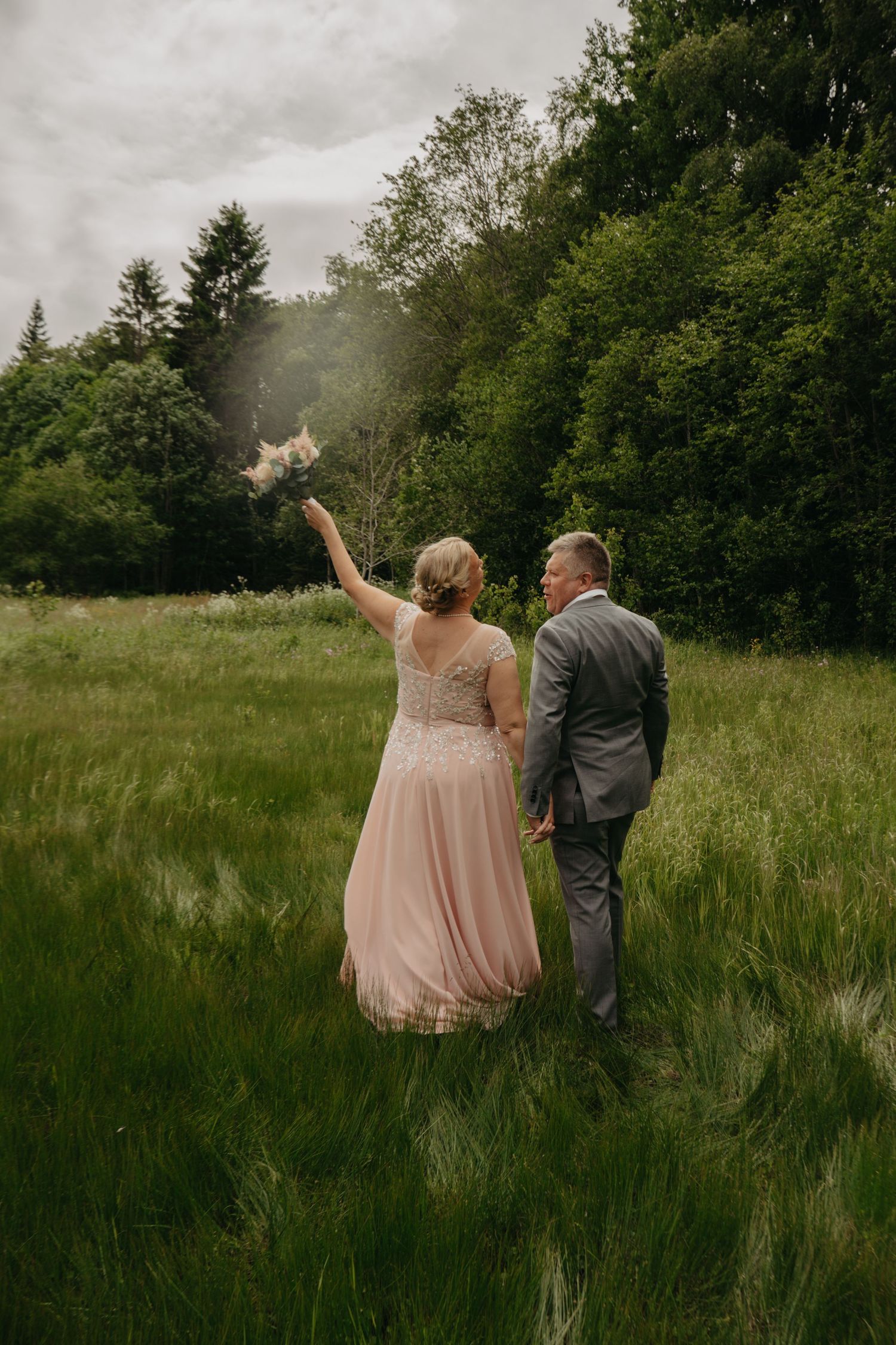 A couple in wedding attire stands in a meadow with the bride tossing a bouquet against a backdrop of evergreen trees.
