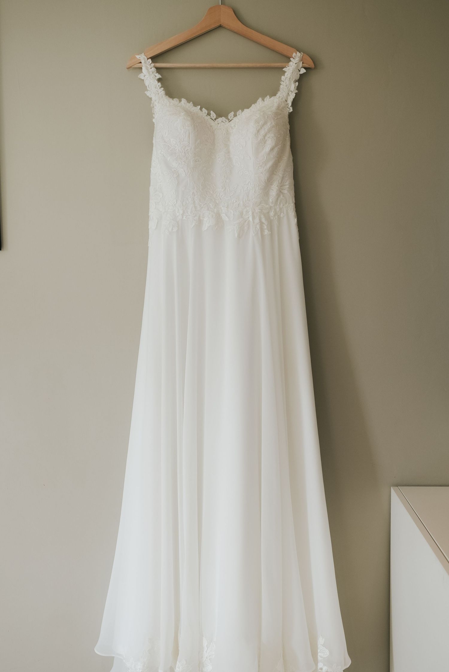 White chiffon wedding dress with delicate lace trim hangs elegantly on a wooden hanger against a neutral wall.