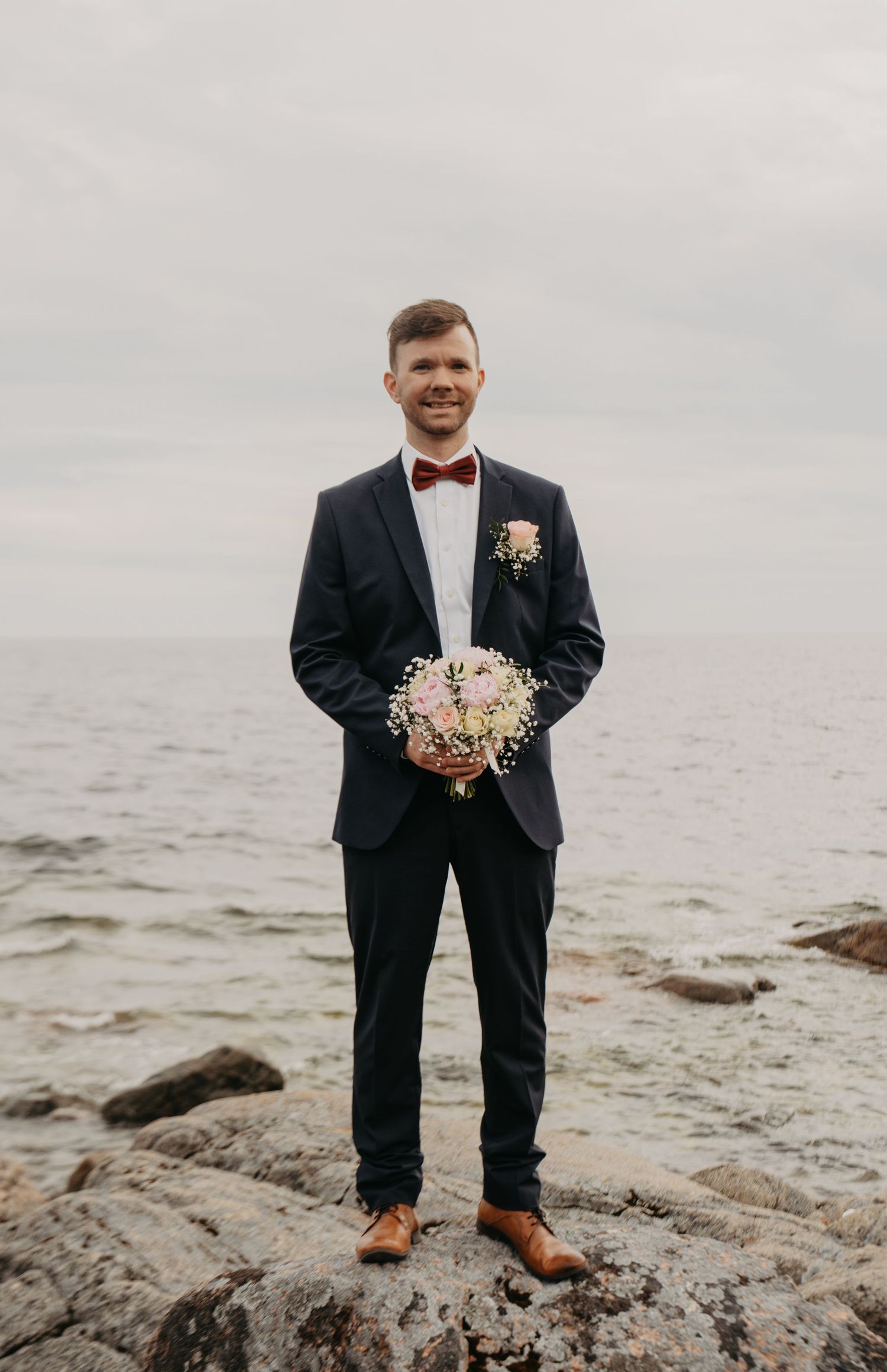 Groom in classic black tuxedo stands on rocky coastal outcrop holding wedding bouquet.