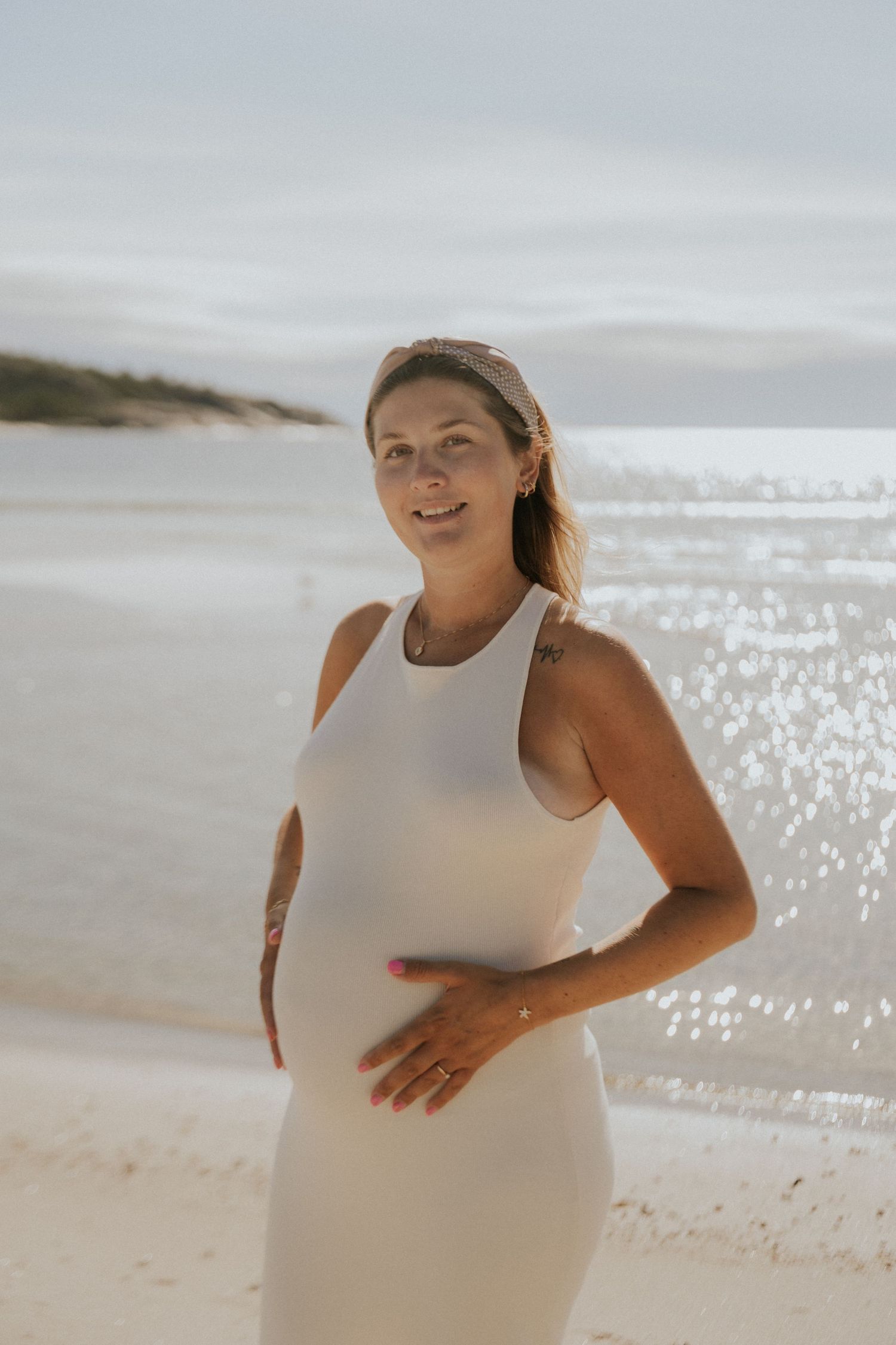 Expectant mother in white dress posing on beach with sparkling ocean in background.