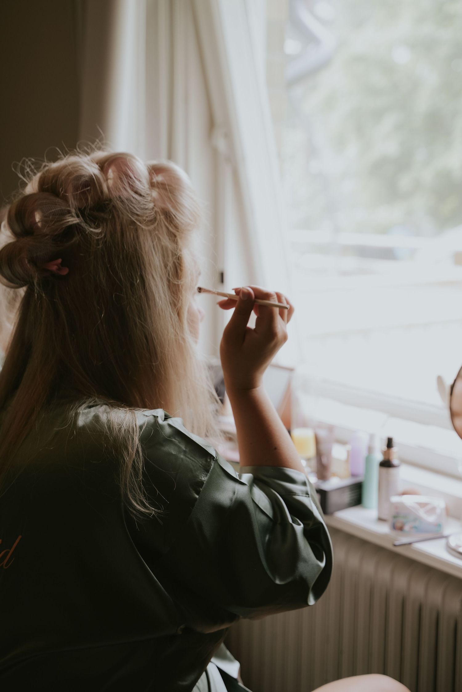A silhouetted figure sits by a window in dim lighting, applying makeup in a sequence of similar poses.