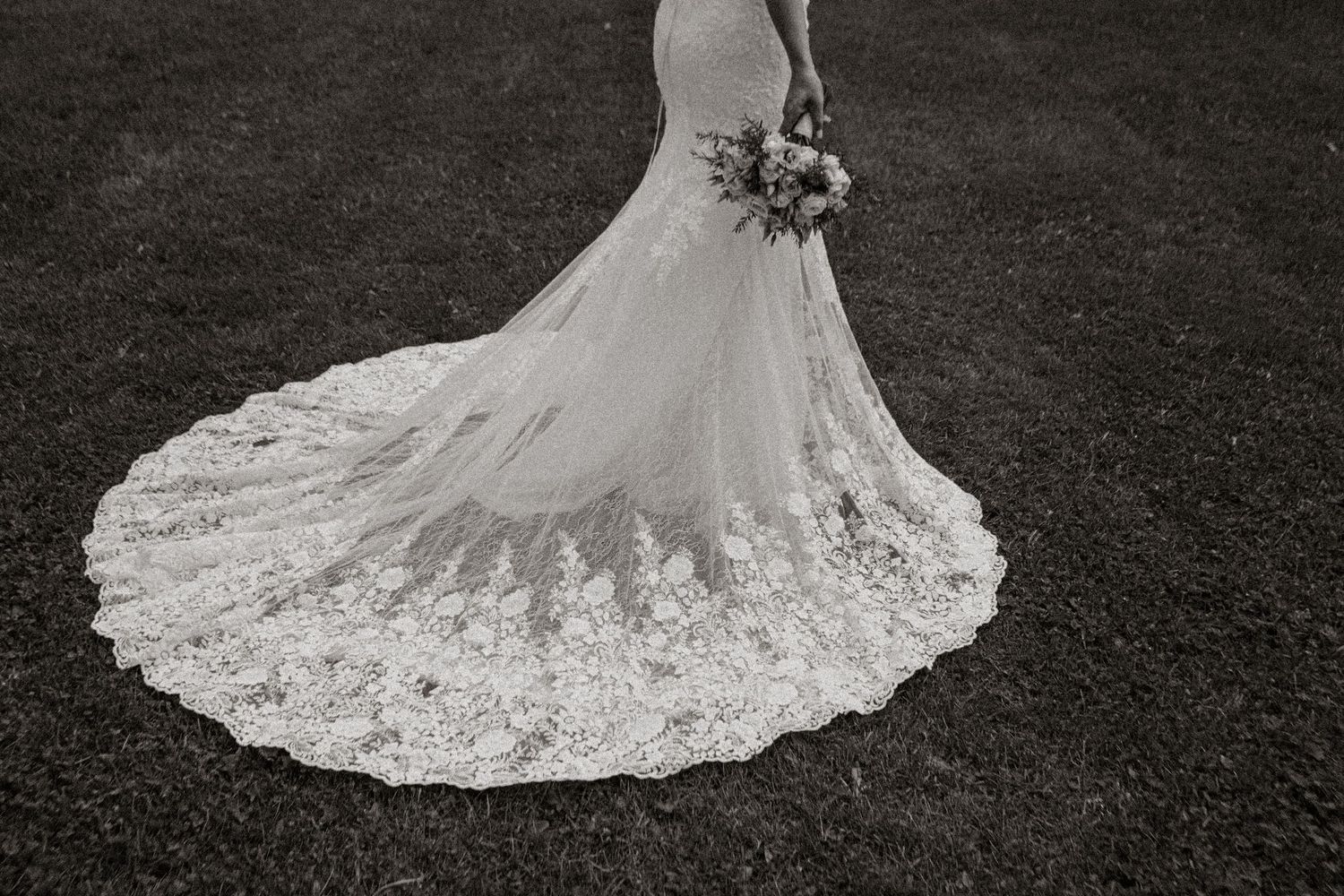 An elegant lace wedding dress train spreads across dark grass in black and white.
