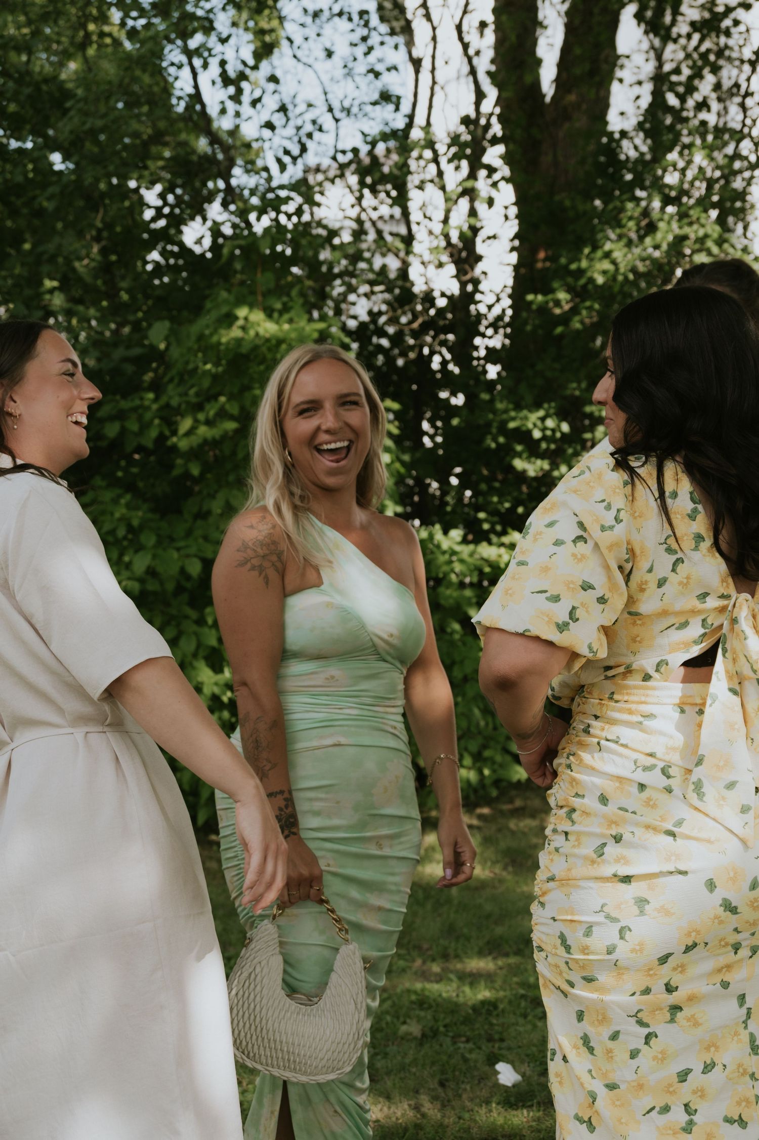 A group of people in formal attire laugh together outdoors during a sunny gathering under trees.