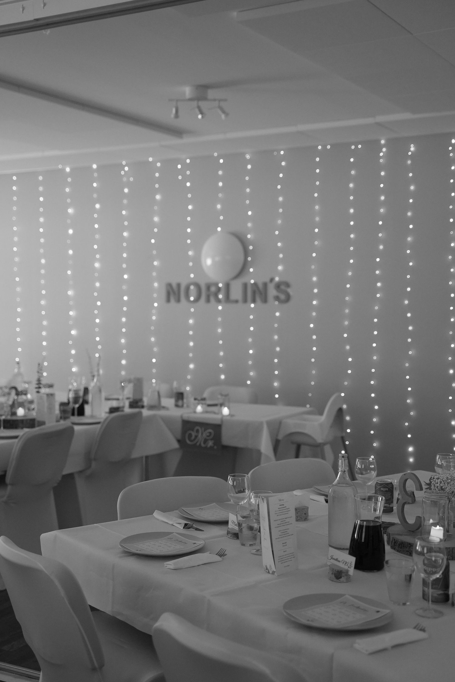 Black and white panoramic view of an elegant dining room setup with string lights and place settings on tables.