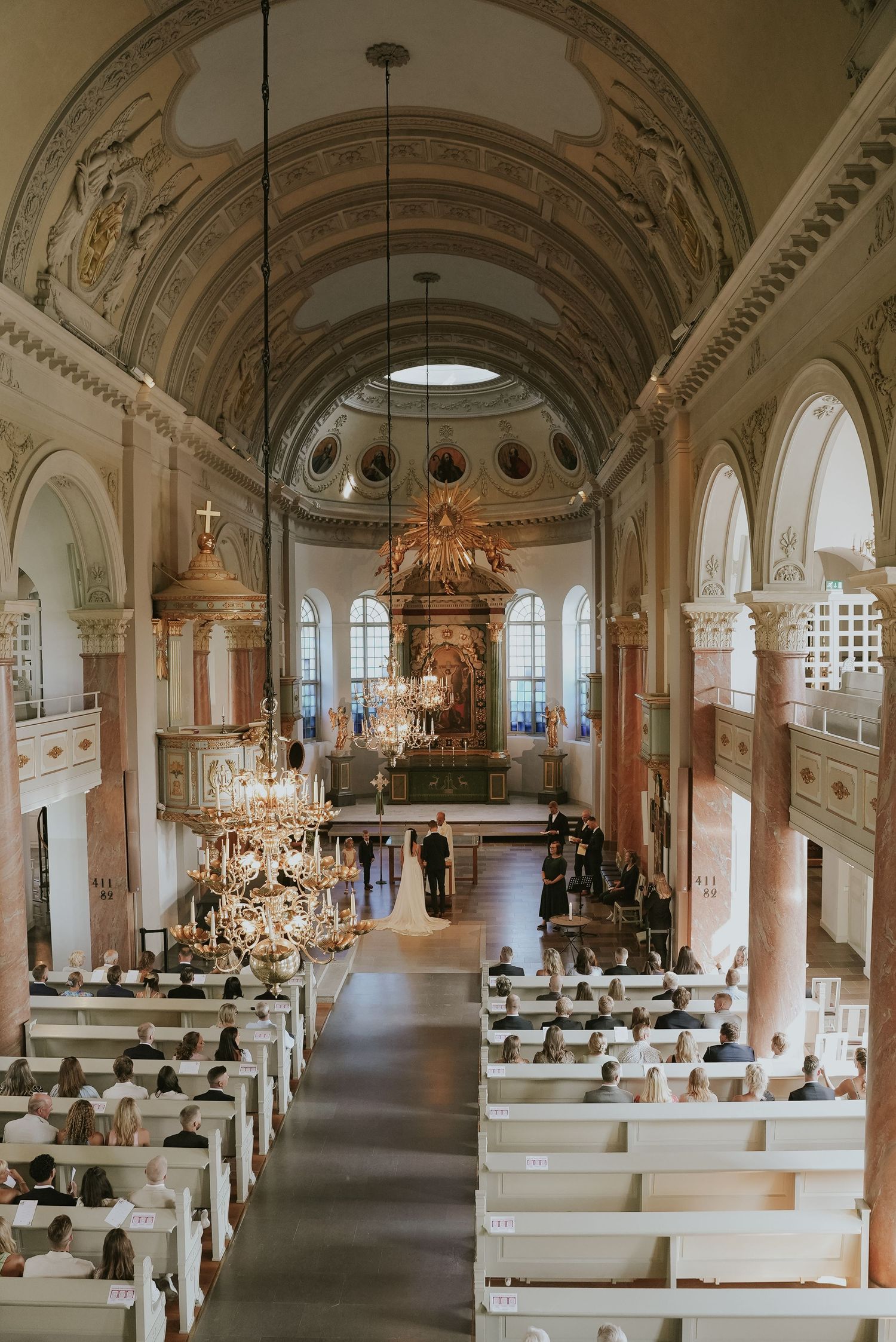 Elegant church interior with white pews, ornate chandeliers, and vaulted ceilings in a panoramic view.