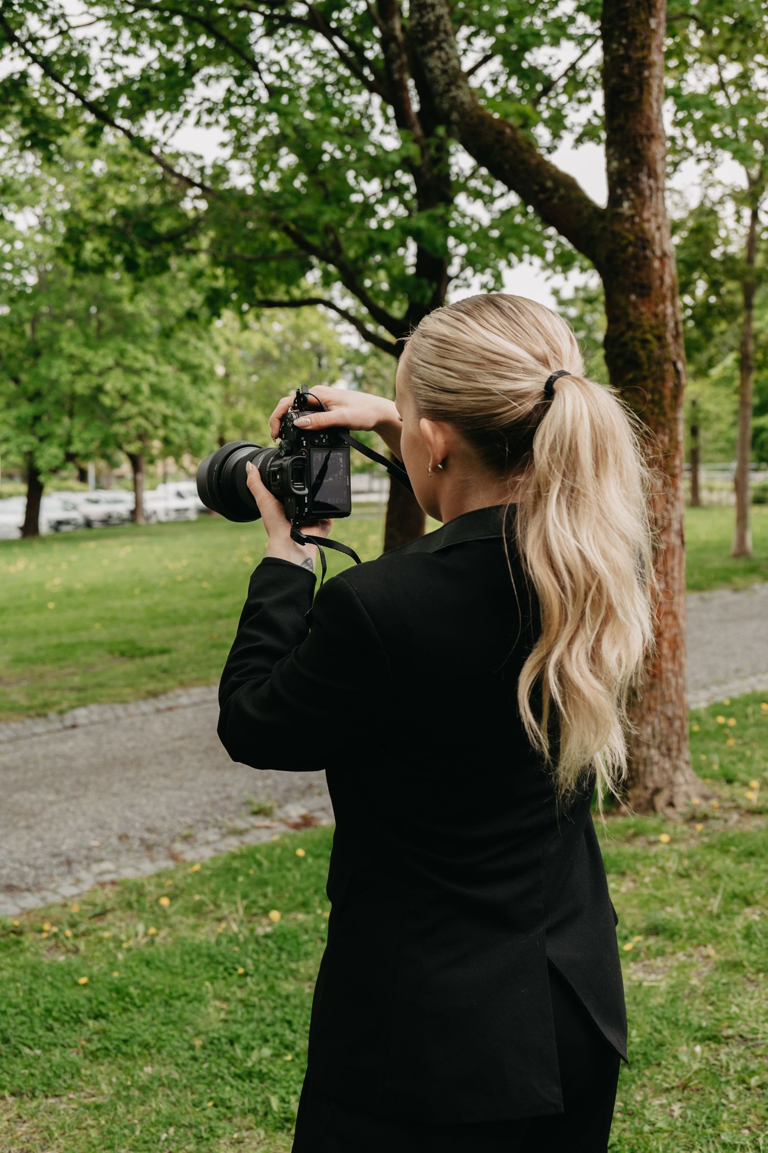 Photographer wearing black jacket takes photos in a green park setting with trees in the background.