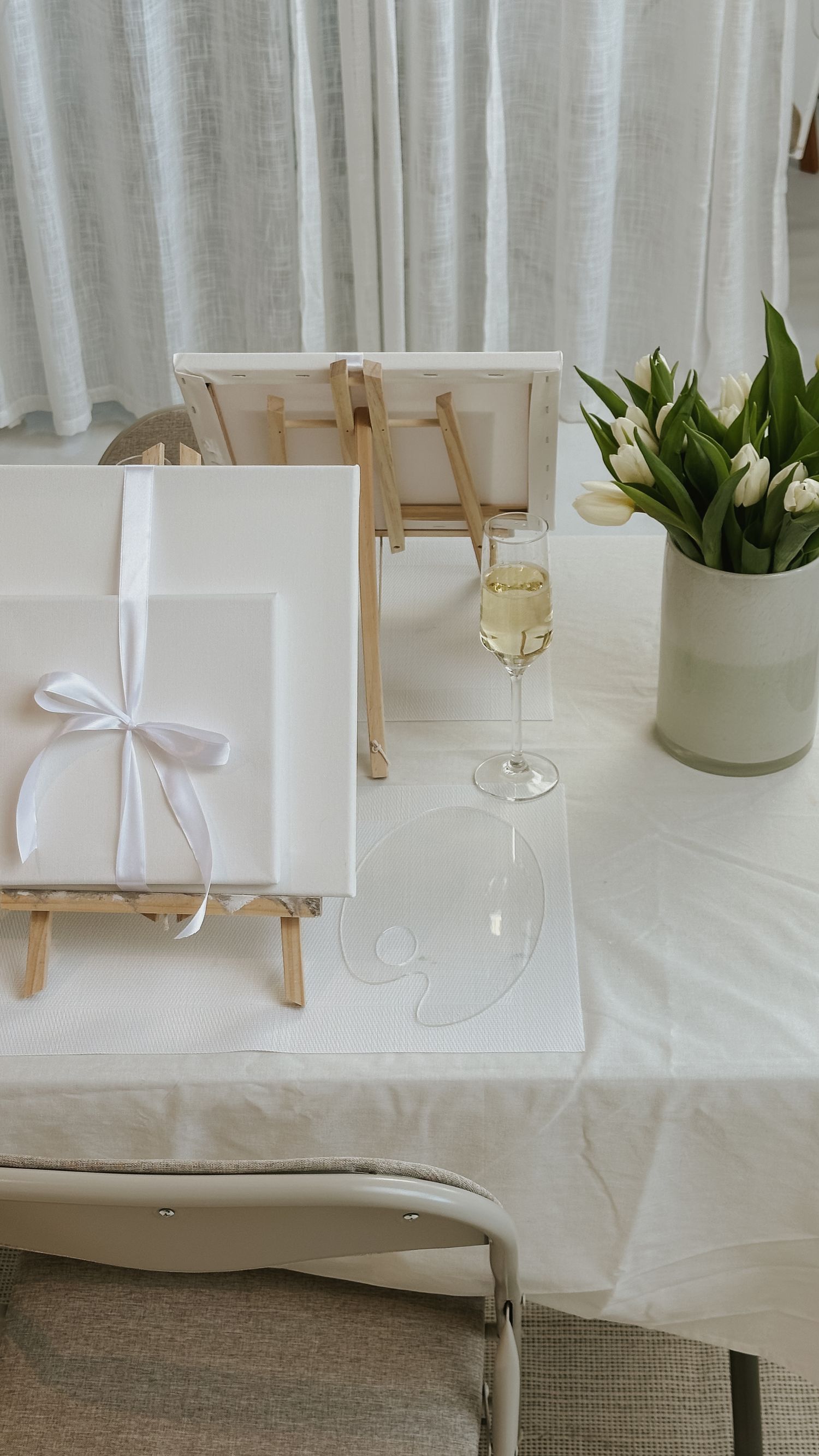 White gift bags with ribbons arranged on a table next to wine glasses and potted plants create an elegant display.