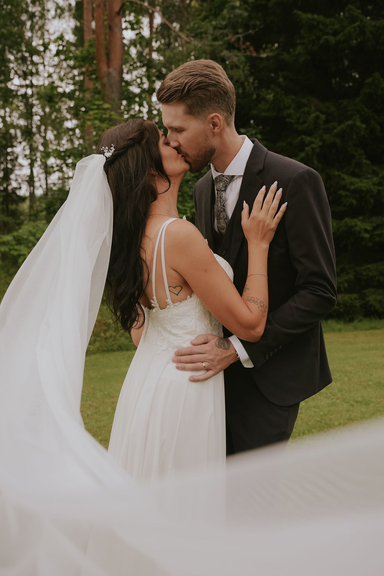 A romantic wedding moment captured outdoors with a flowing white veil and elegant attire against a forest backdrop.