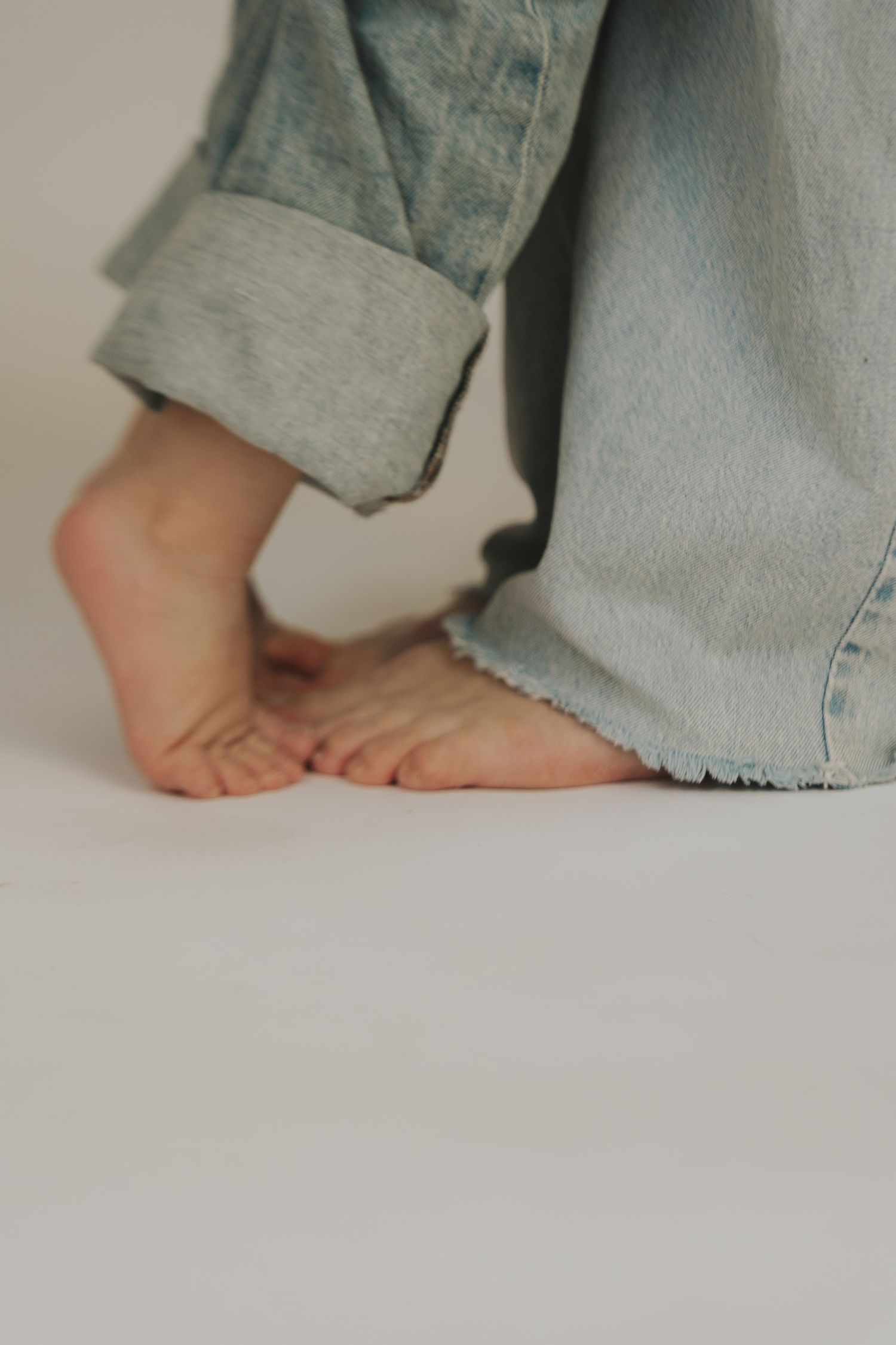 Bare feet peeking out from under light blue denim jeans against a neutral background.