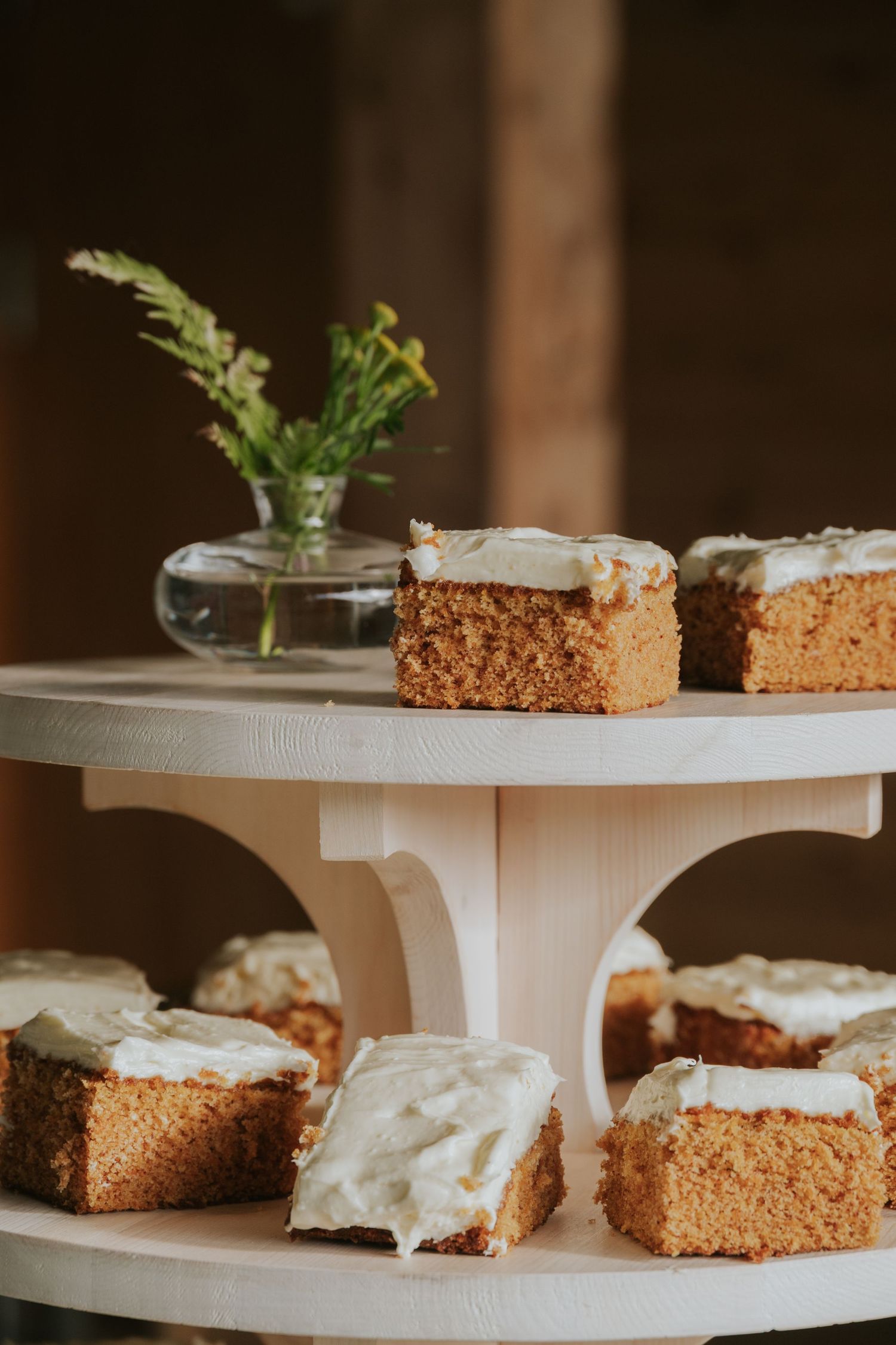 Elegant dessert display featuring frosted carrot cake squares garnished with fresh herbs on white tiered serving stands.