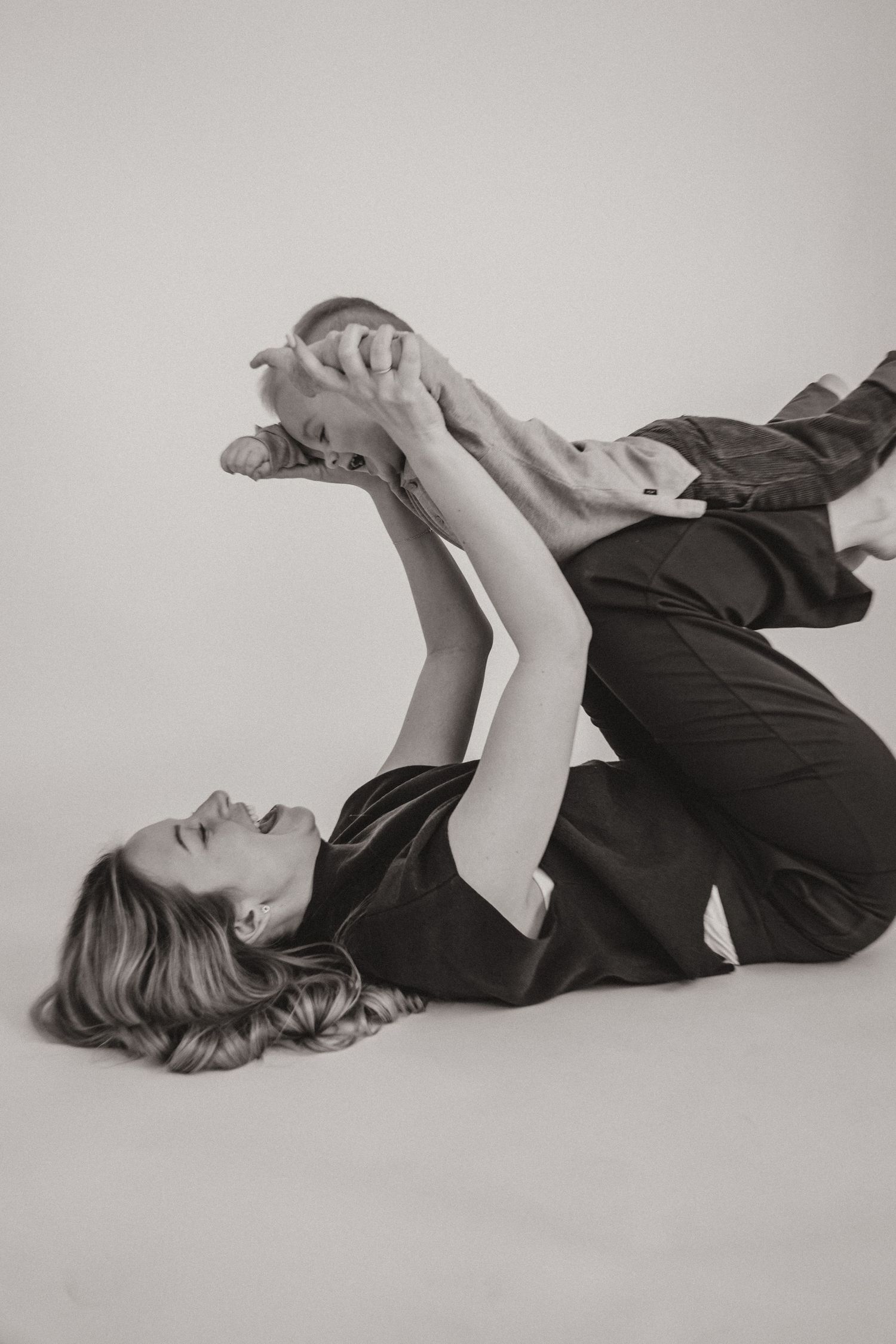 Black and white artistic portrait of person lying down holding a cat in playful pose against white background.