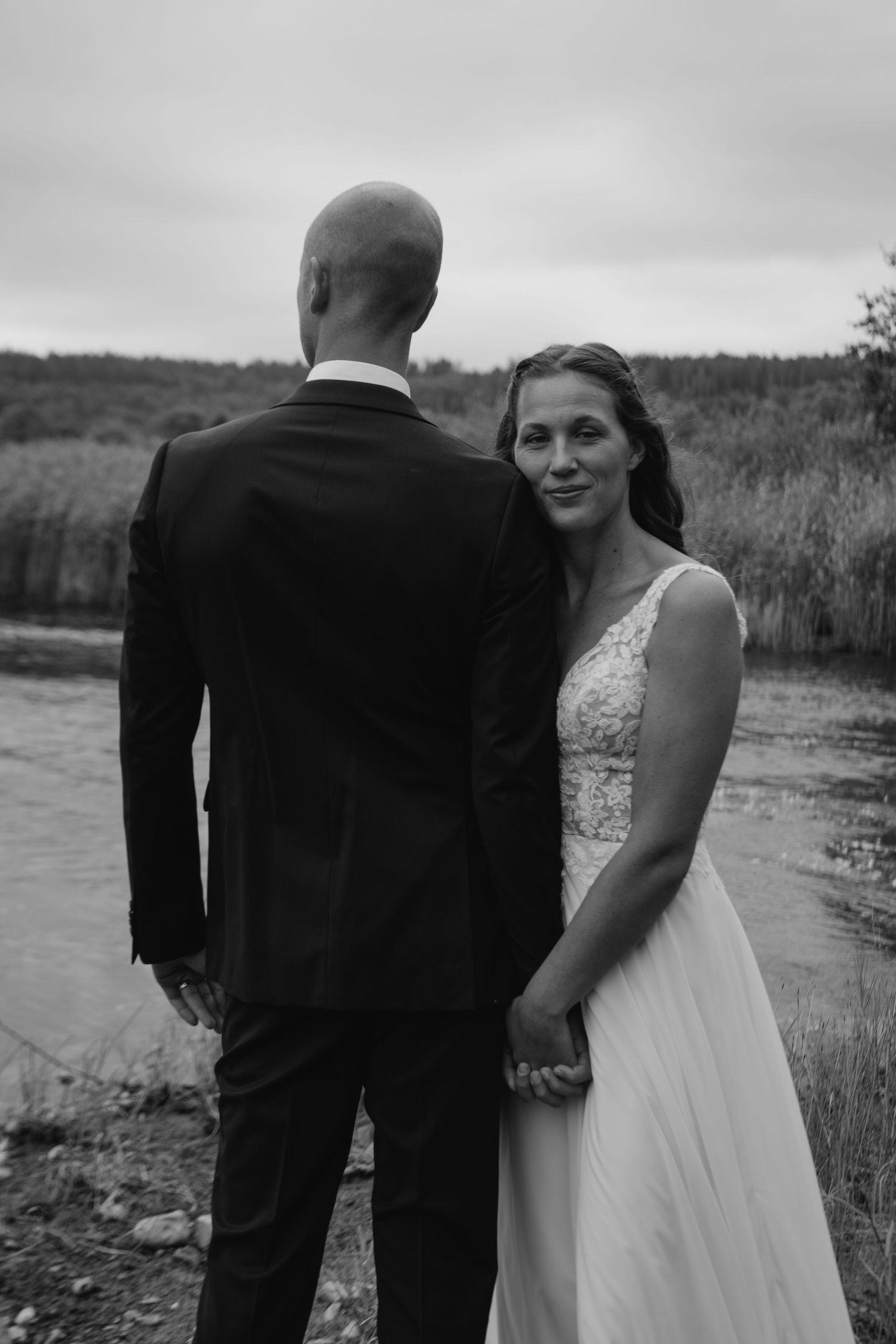 Black and white portrait of newlyweds standing by lake in elegant attire.