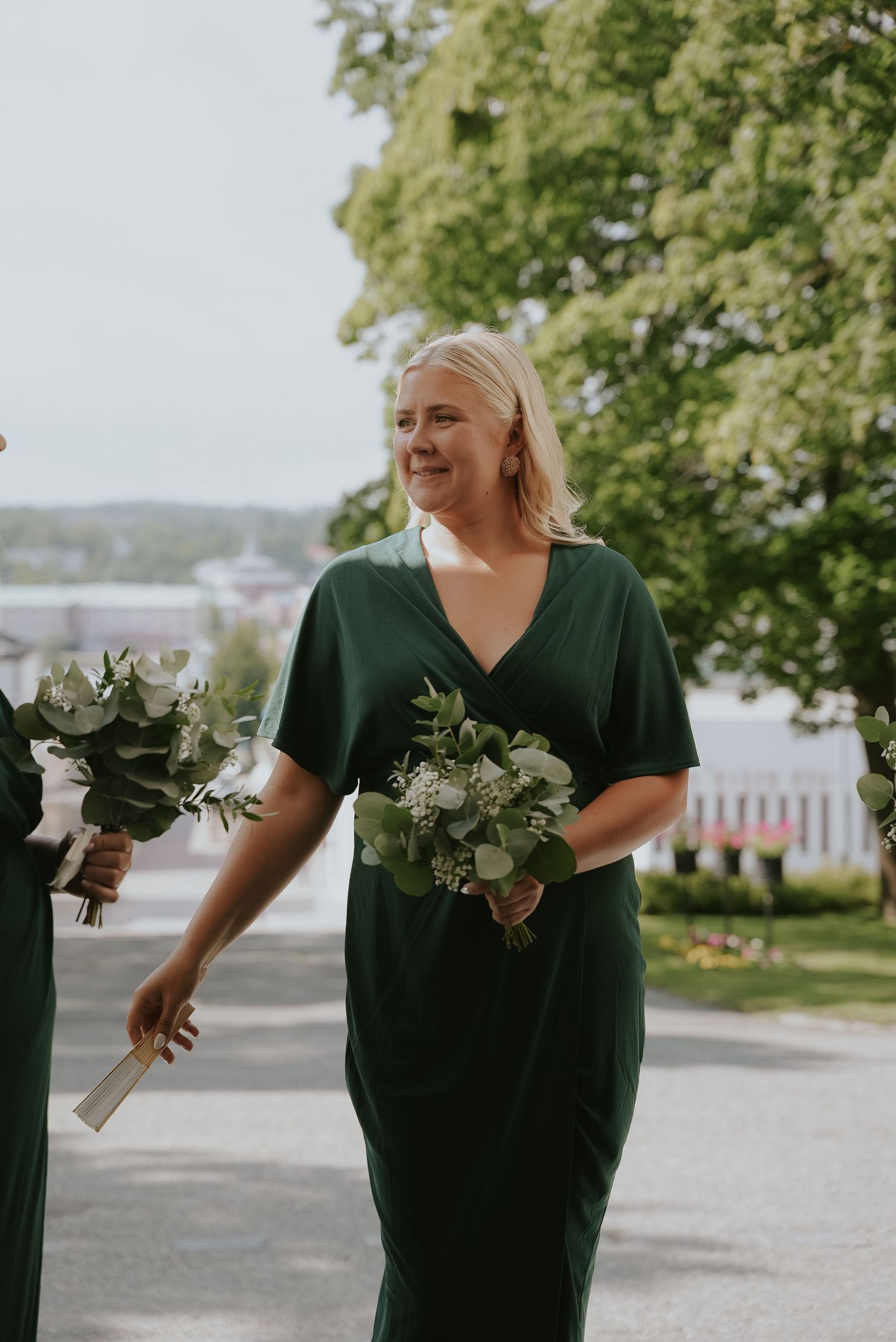 Bridesmaid in emerald green dress carries white floral bouquet while walking outdoors on wedding day.