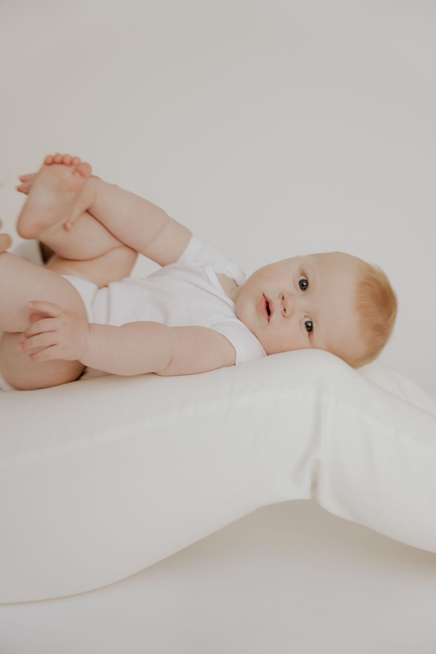 Baby in white onesie lying on soft white surface looking at camera.