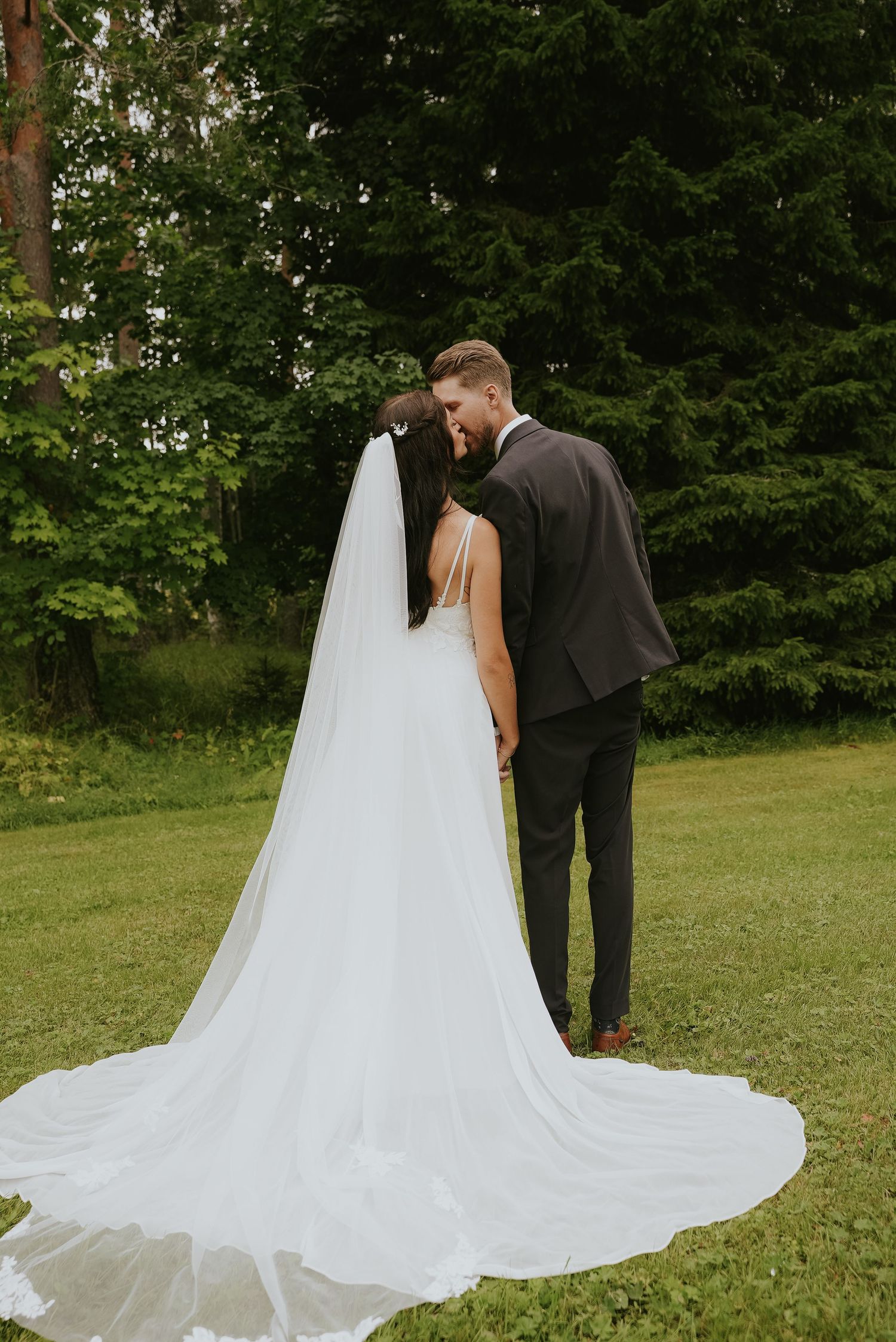Wedding couple in elegant attire posing on grassy lawn with flowing cathedral veil.