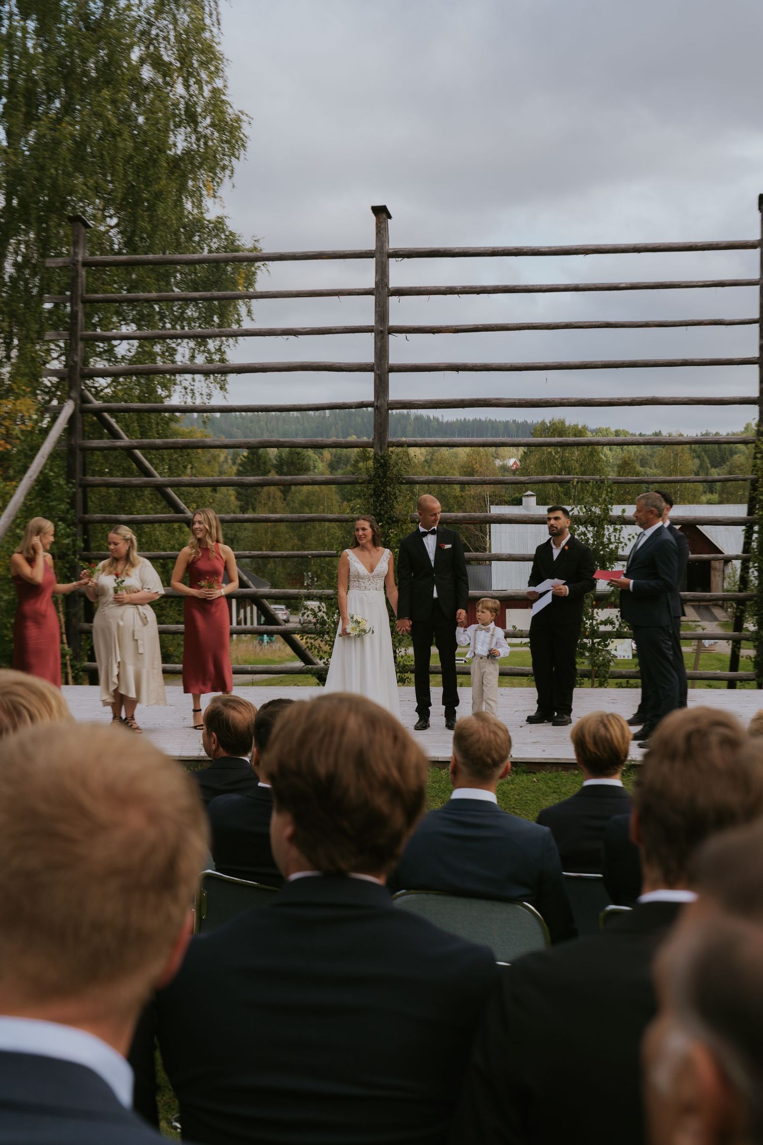 Wedding party stands in line during outdoor ceremony with wooden fence backdrop.