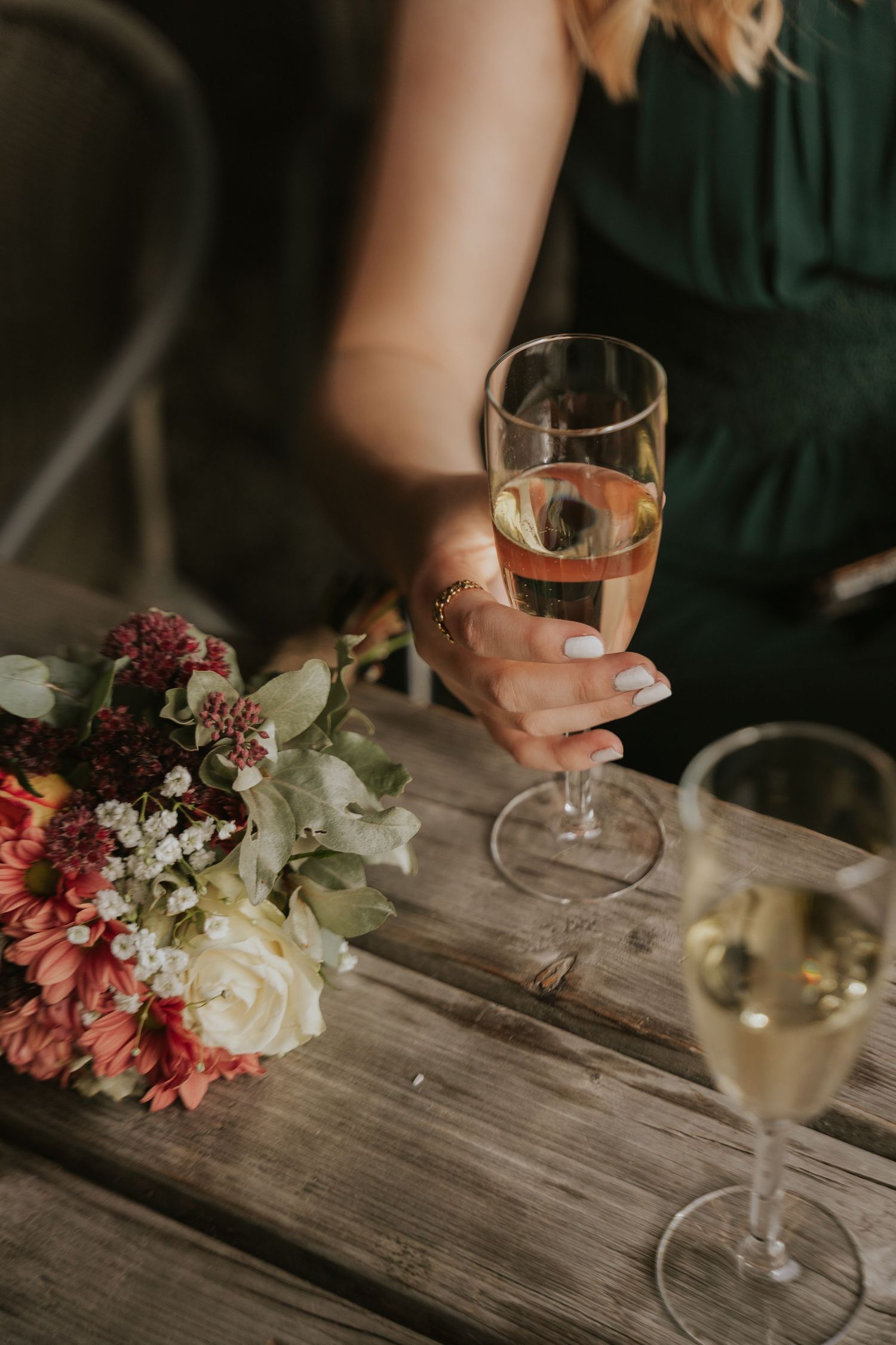 Wine glasses and pink floral arrangements sit on a rustic wooden table during an intimate gathering.