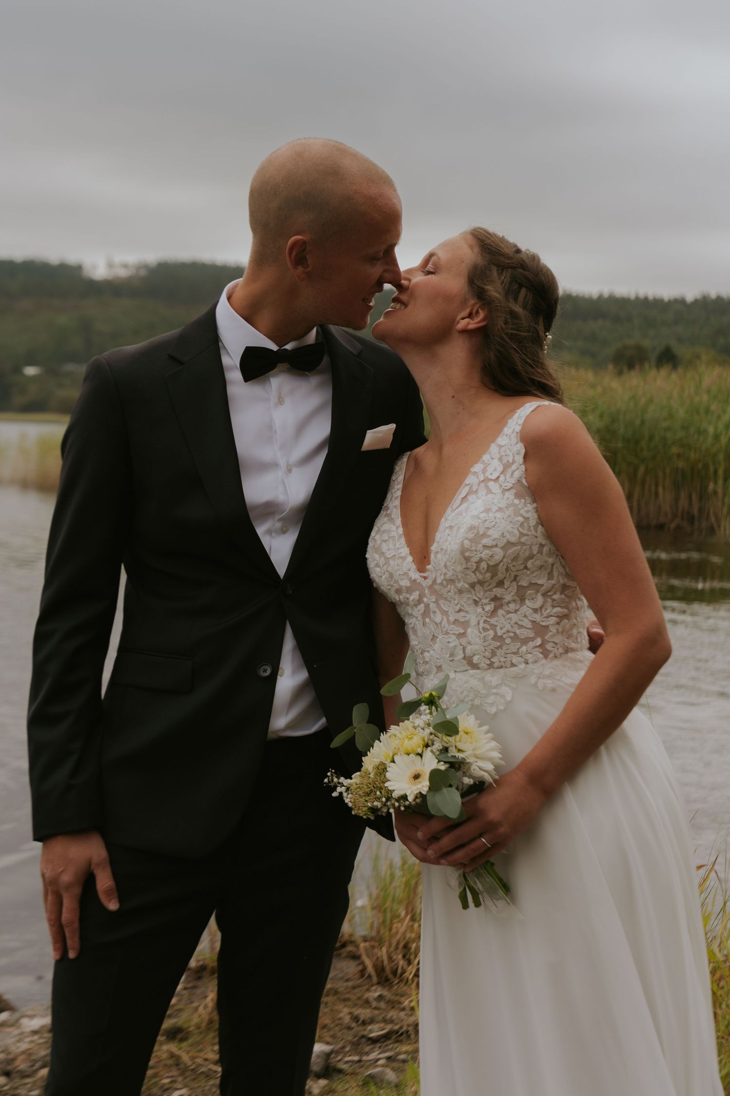 Romantic wedding couple sharing intimate moment by lake with cloudy sky backdrop.
