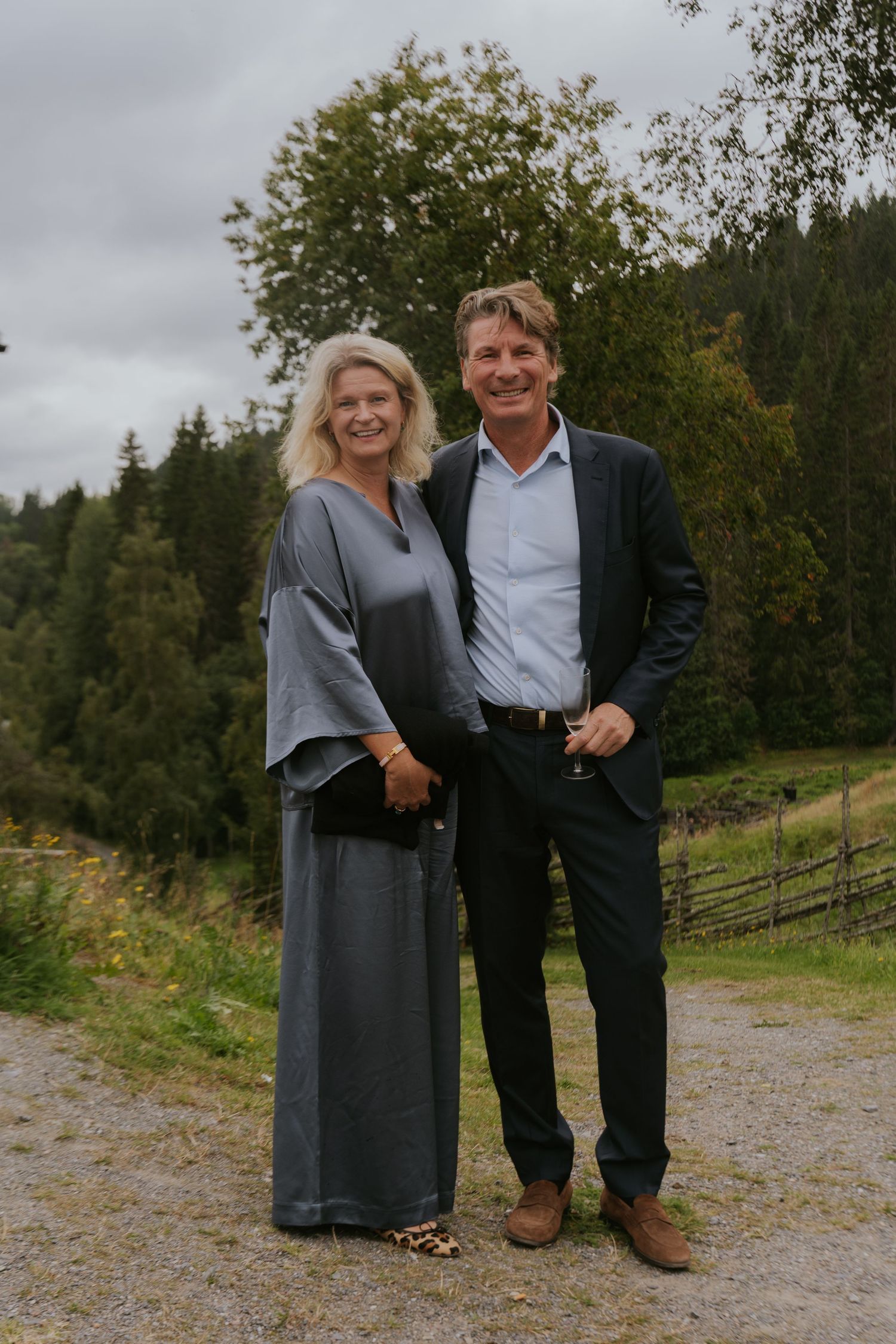 Couple in formal attire poses together on gravel path with autumn trees in background.