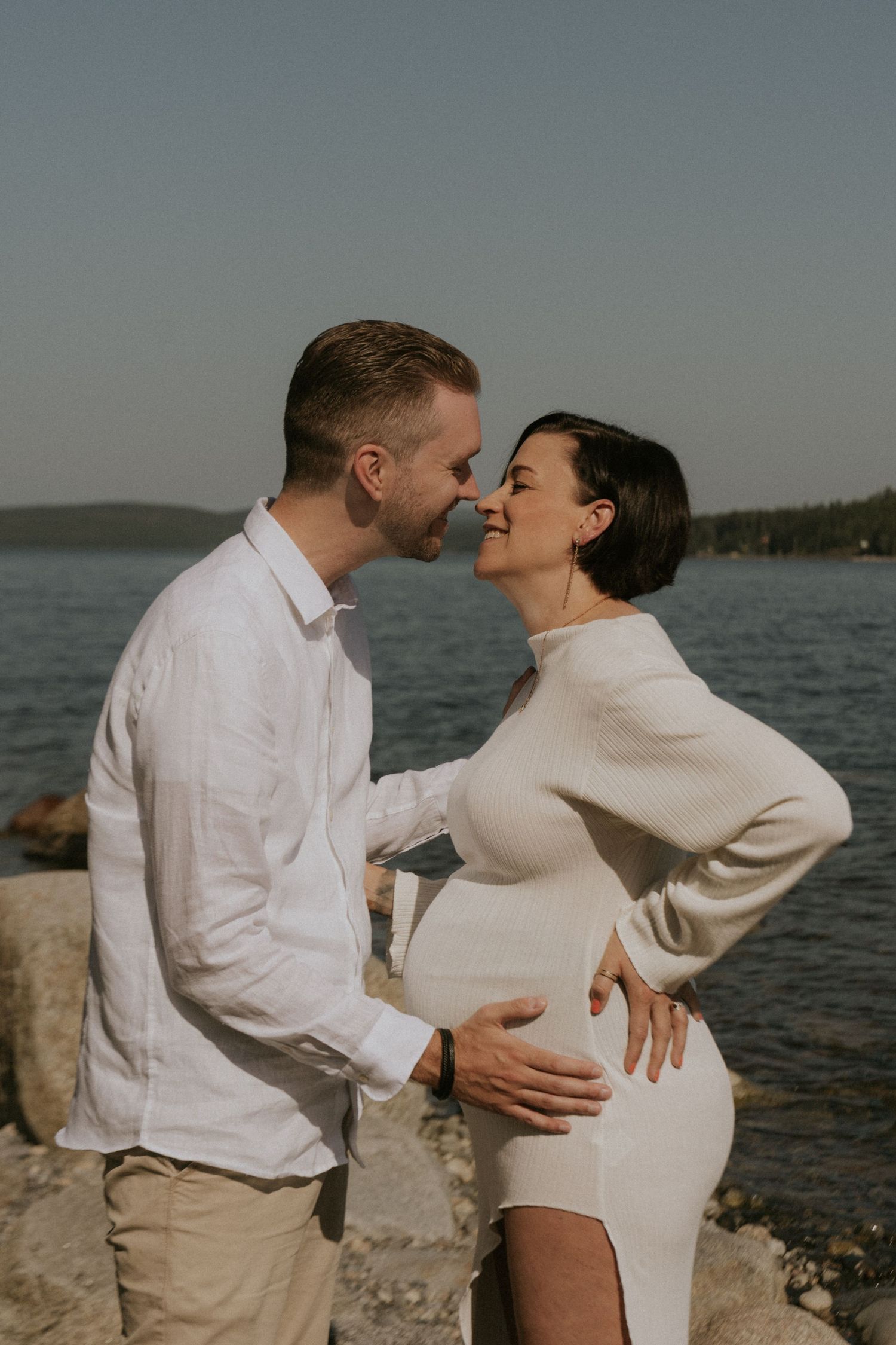 A romantic couple in white clothing embraces and laughs together by the waterfront.