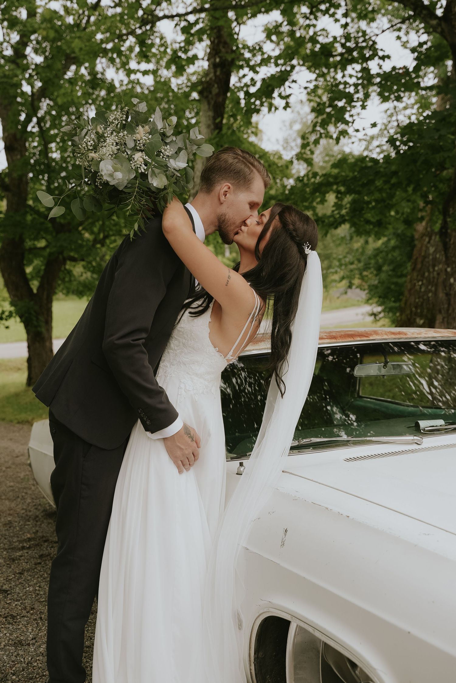 A romantic wedding moment captured next to a white vintage car under lush green trees.
