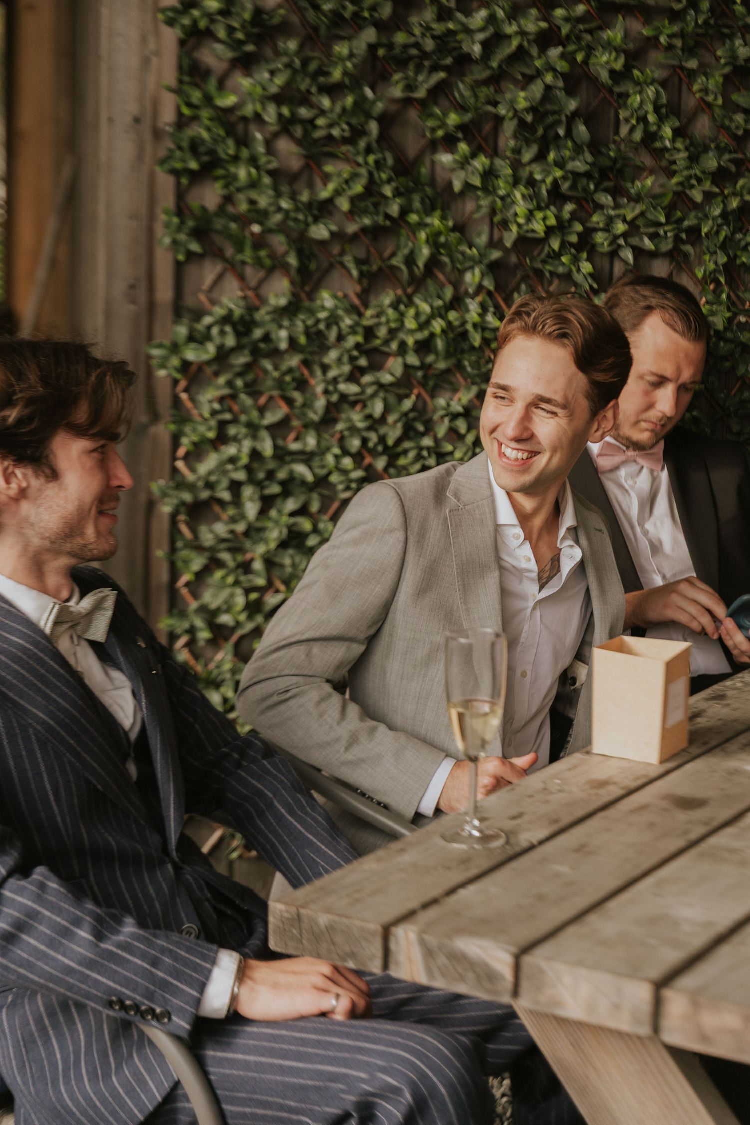 Three friends in suits share drinks and laughter at an outdoor table with ivy-covered wall behind them.