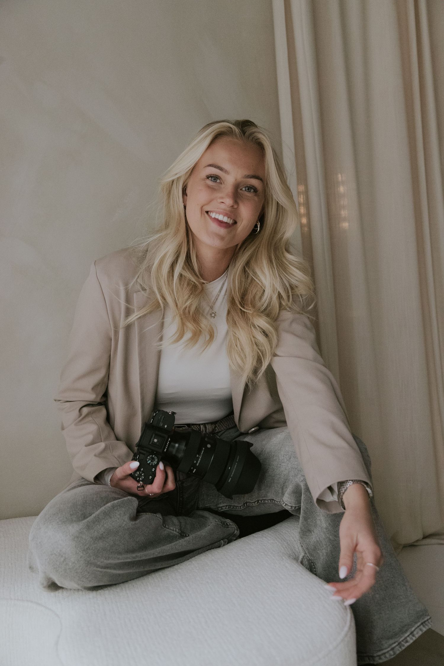 A person in a beige blazer and white top lies on a white couch holding a camera while smiling.