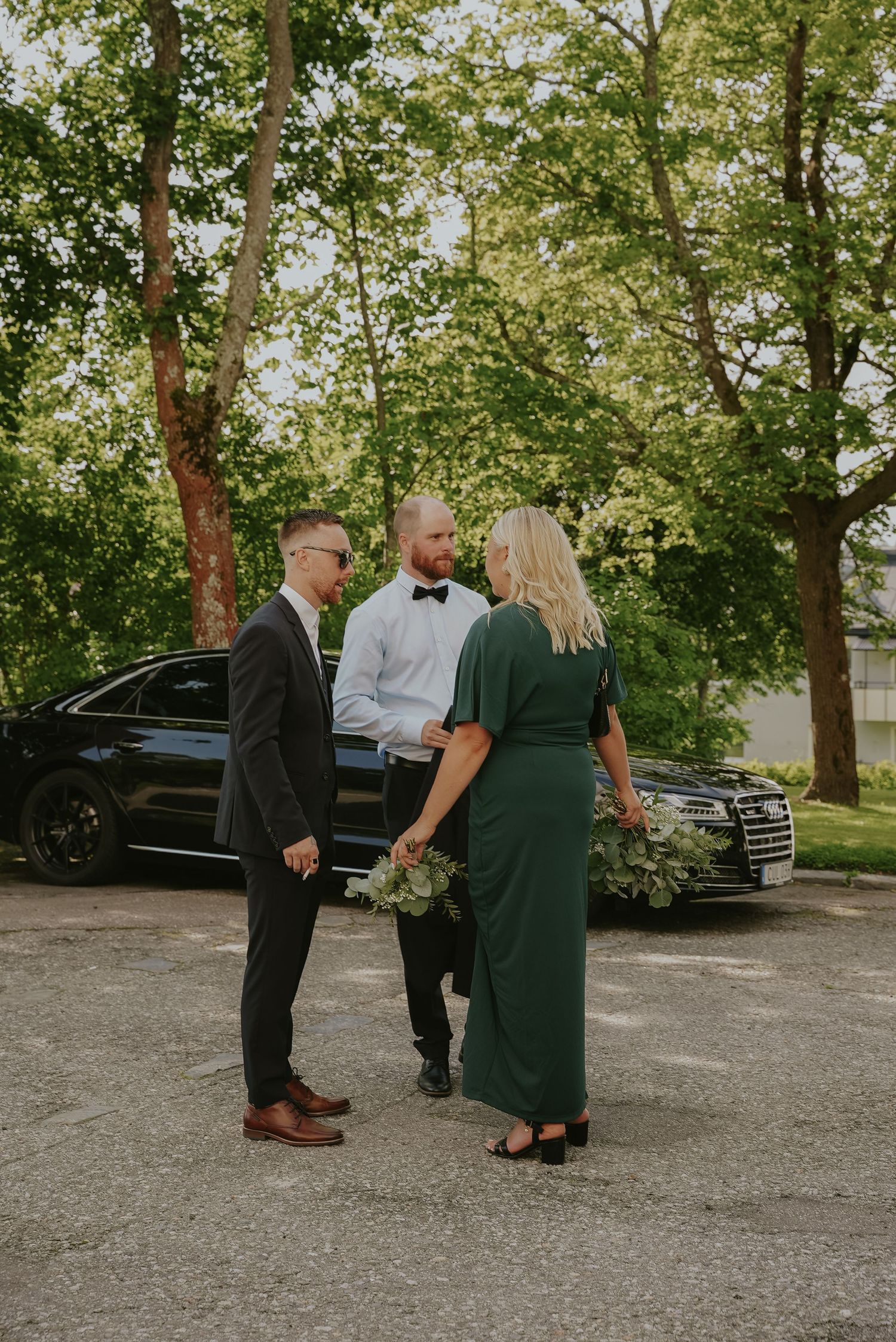 Three people stand in formal attire near a luxury car on a tree-lined driveway during a special occasion.
