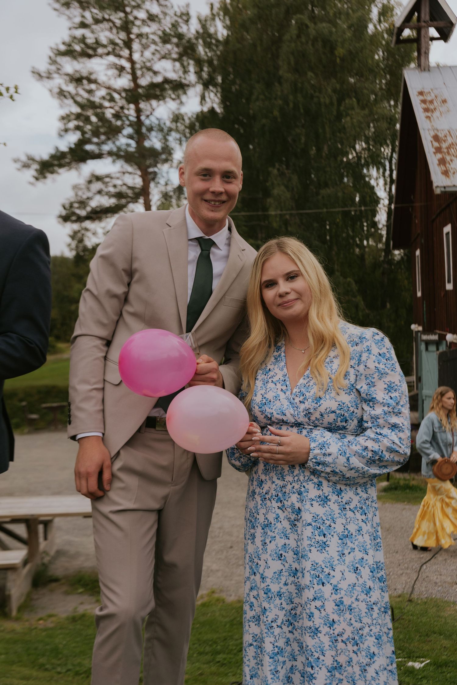 Wedding guests in floral dress and beige suit pose with pink balloons at outdoor venue.