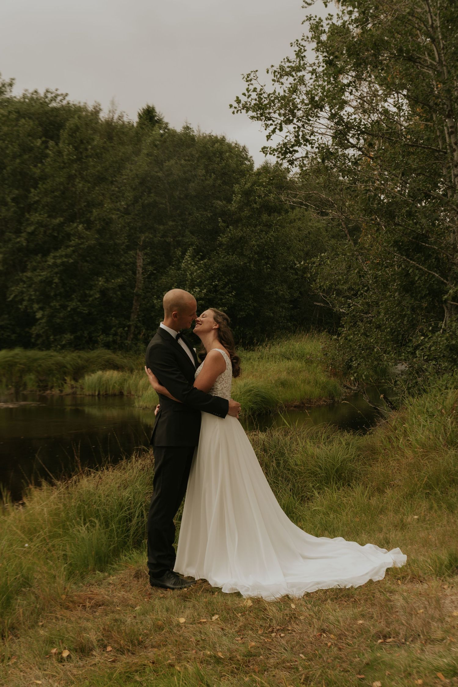 A romantic wedding couple embraces by a serene pond surrounded by lush greenery.