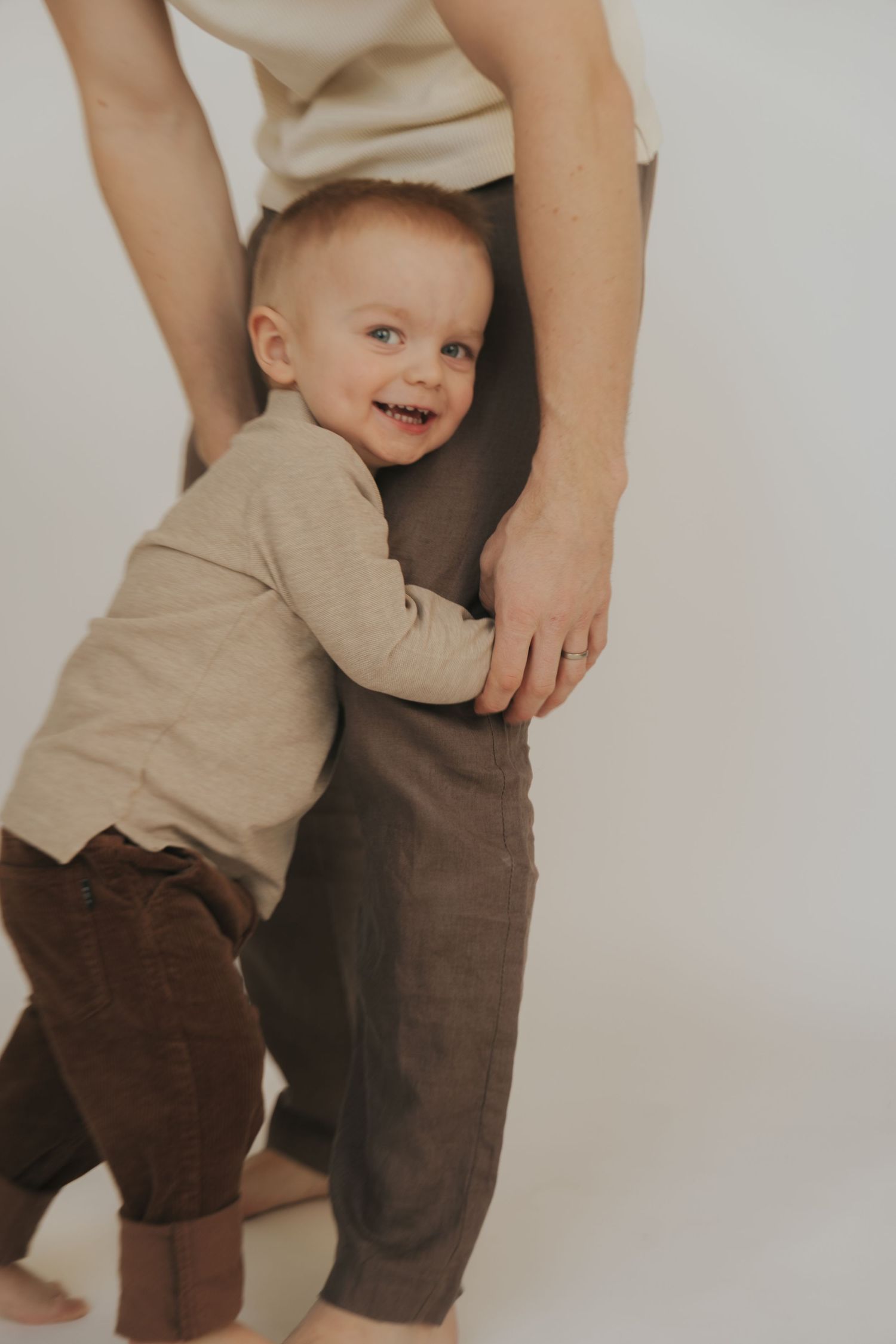 A small child in beige clothing holds onto an adult wearing brown pants while learning to walk against a white background.
