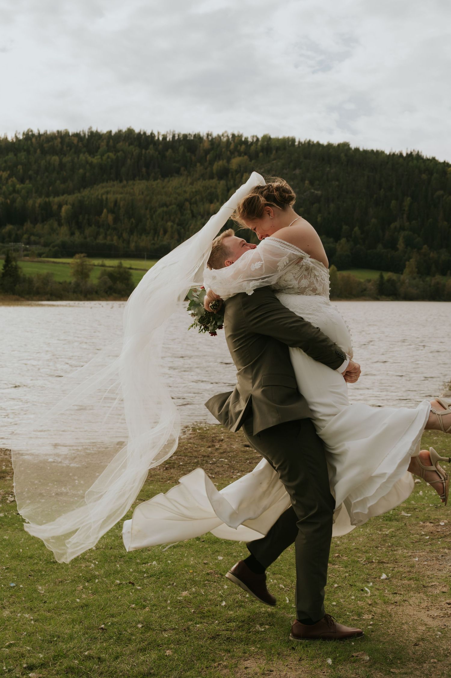 Wedding couple playfully runs with flowing veil by a scenic lake.