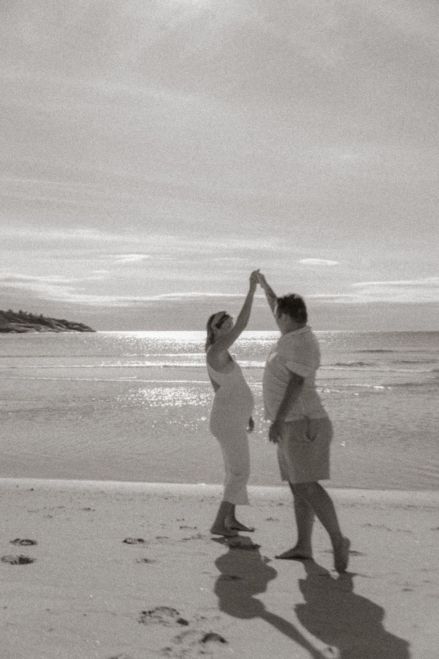 Black and white photo of two people dancing on beach with long shadows.