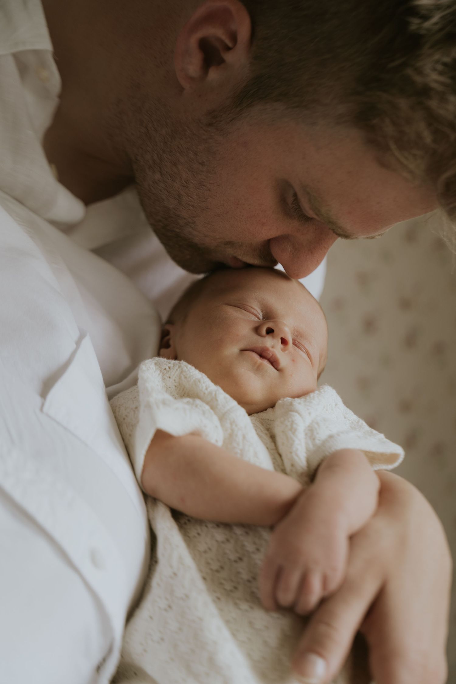 A tender moment between parent and newborn baby in soft white bedding with gentle natural lighting.