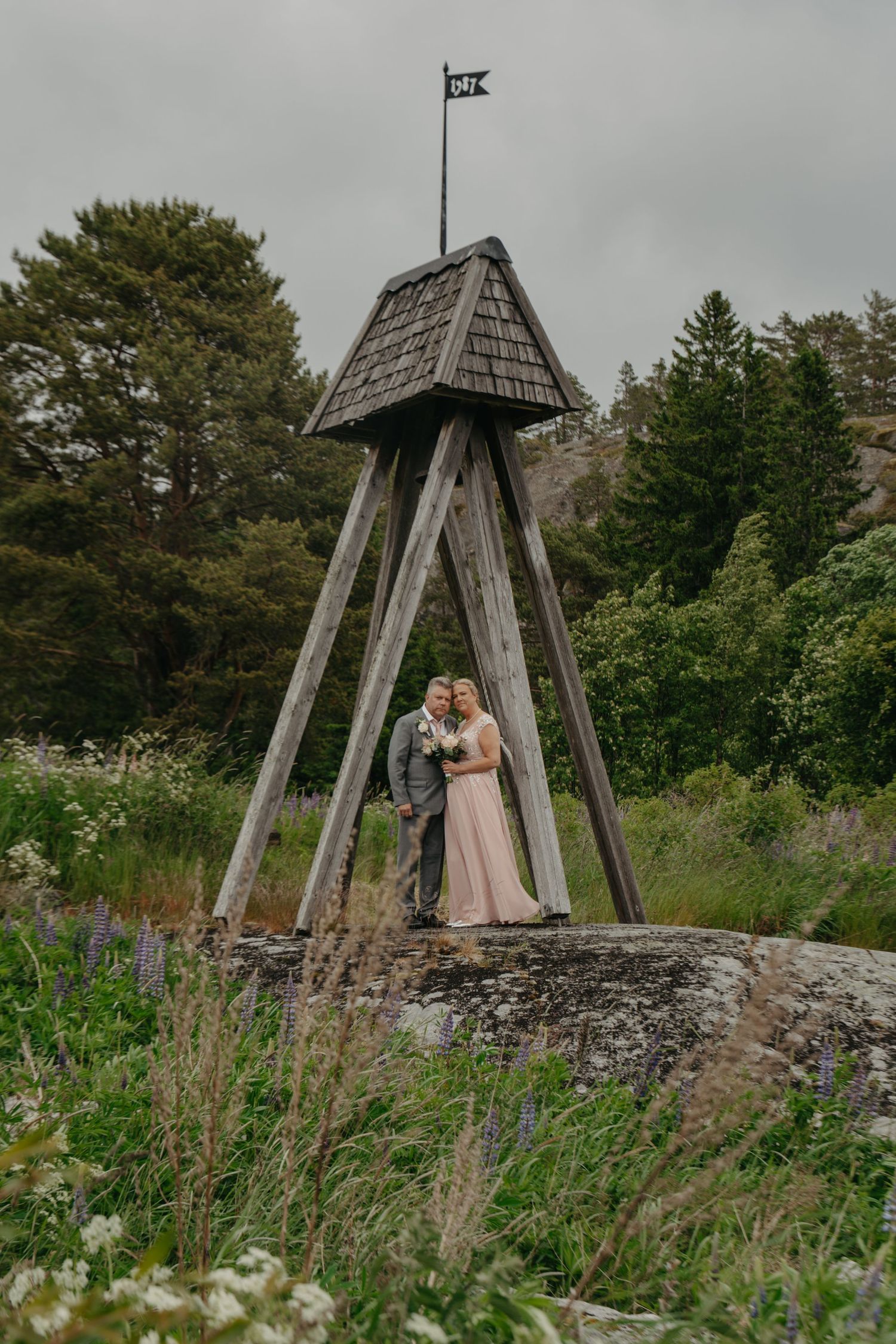 Wedding couple stands under rustic wooden tower structure surrounded by wildflowers.