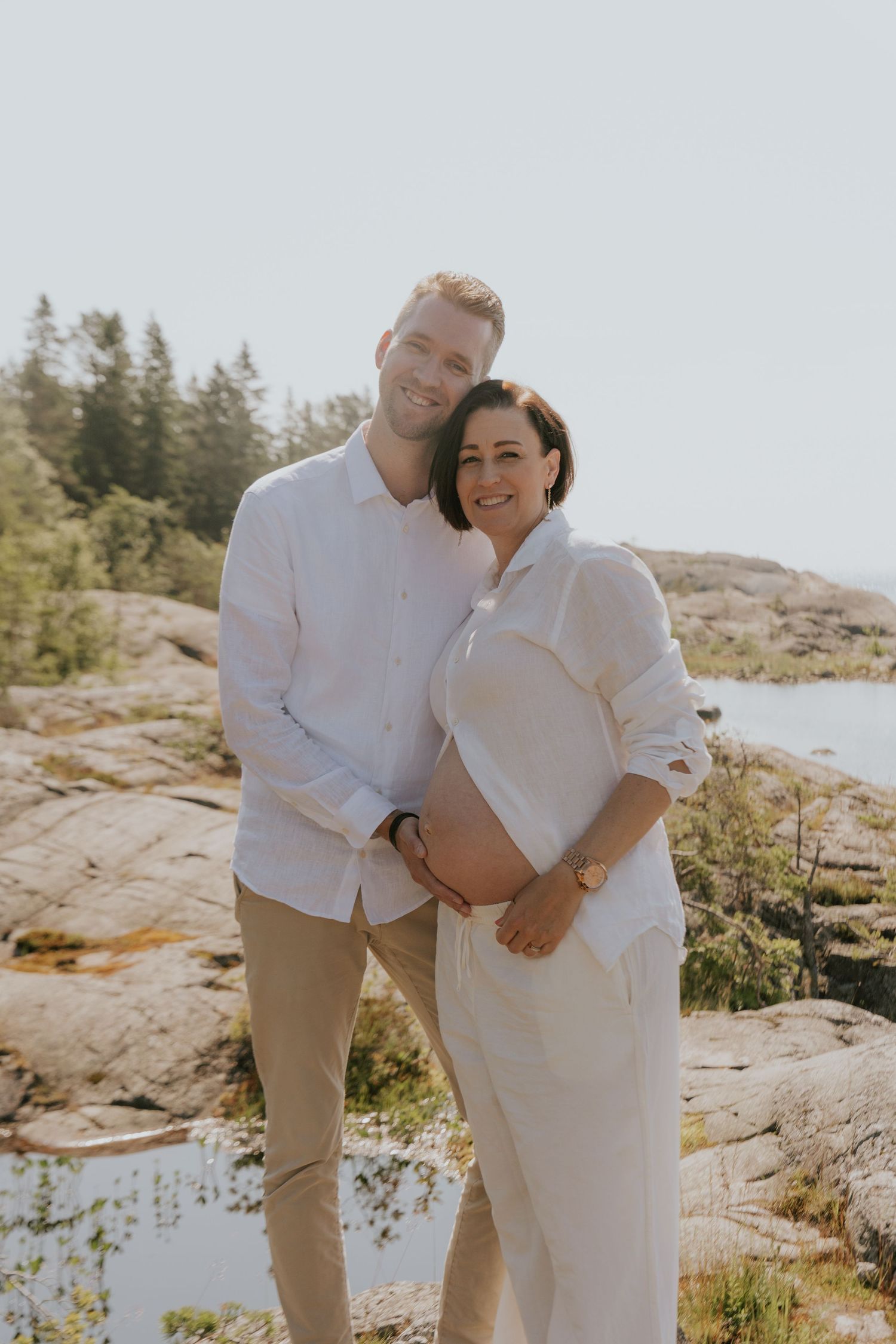 A couple in coordinated white outfits poses lovingly together on coastal rocks with water in background.
