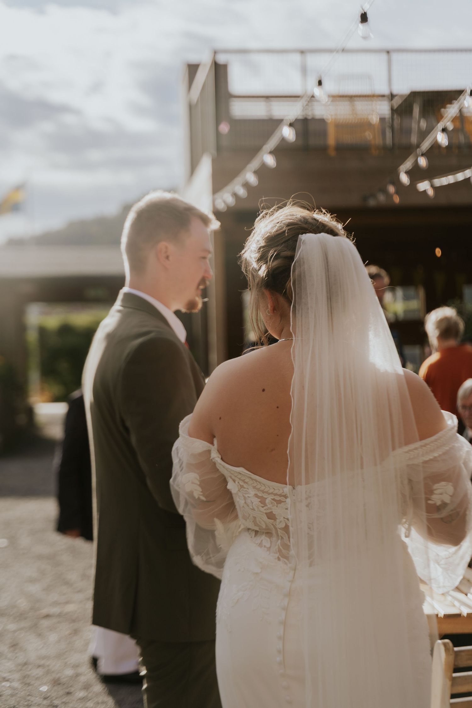 A wedding couple stands together at an outdoor venue with string lights and cloudy skies in the background.